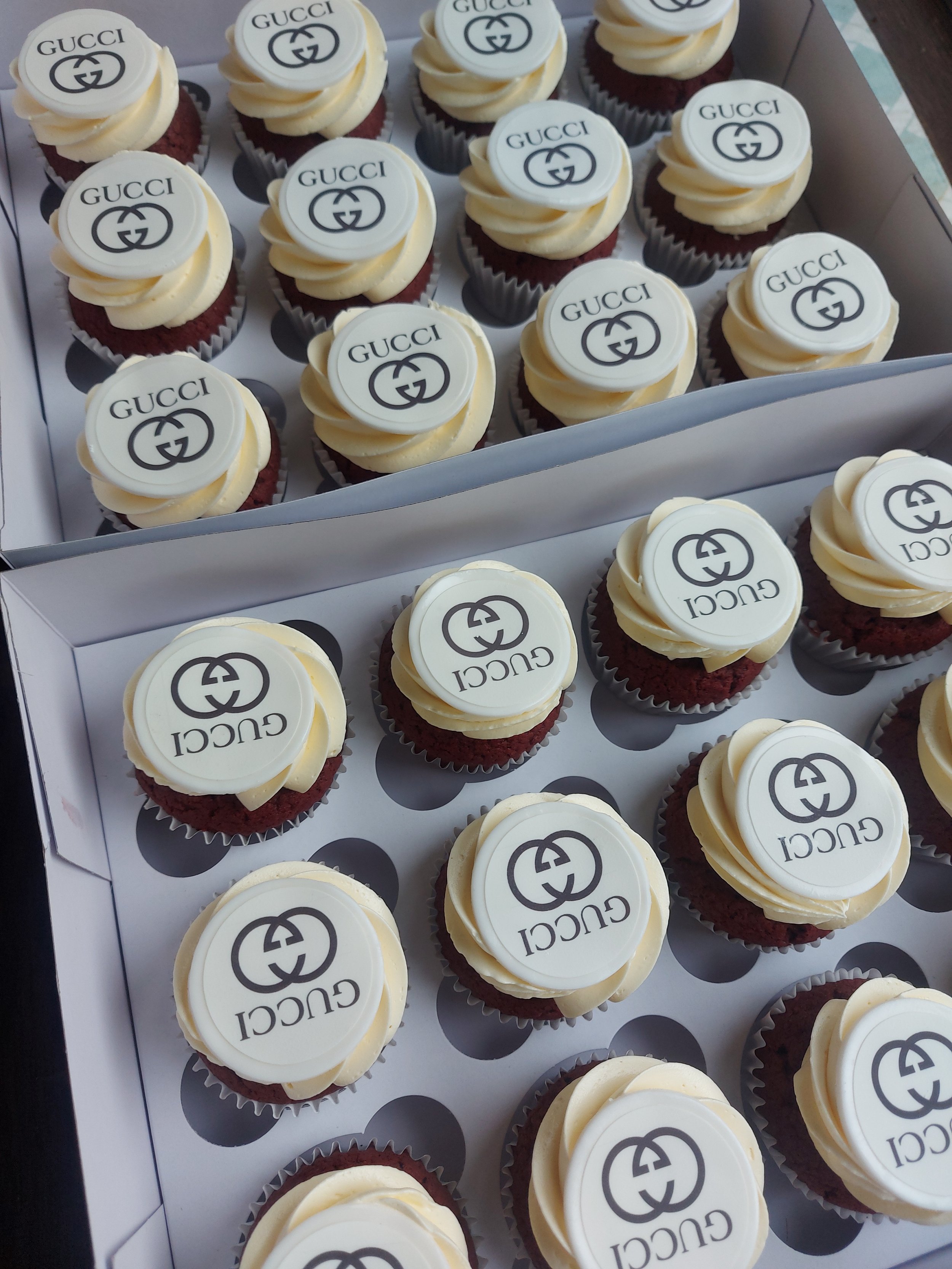 A box of cupcakes decorated with white fondant toppers displaying the Gucci logo in black, topped with swirls of white frosting.