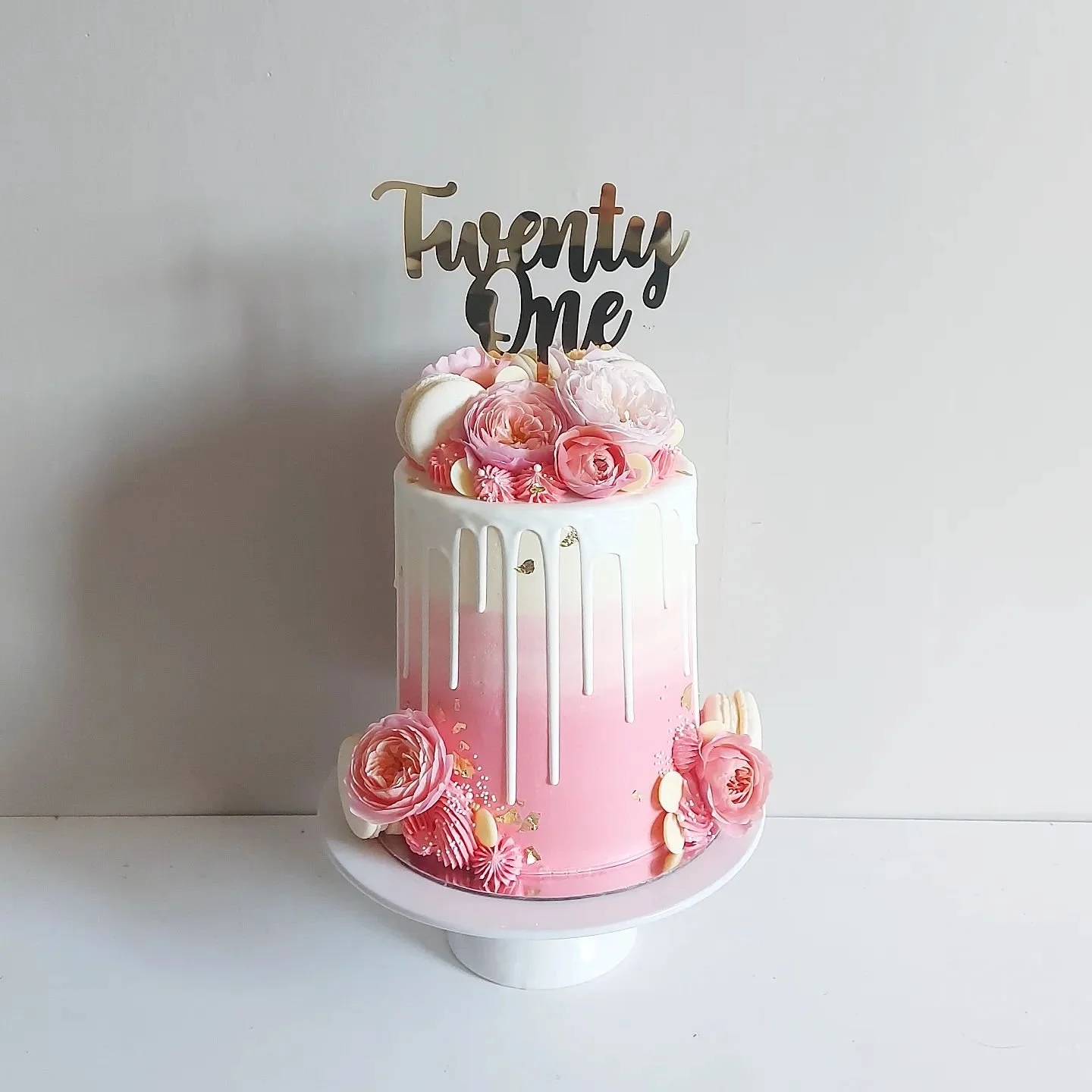 Pink and white tiered cake decorated with pink roses, white chocolate pieces, and gold leaf, topped with a cake topper that says 'Twenty One' in black and gold, against a plain white background.