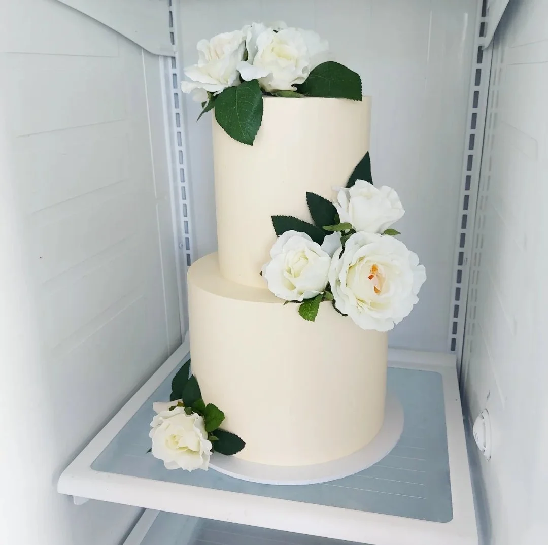 Two-tiered white wedding cake decorated with white roses and green leaves, placed inside a refrigerator.