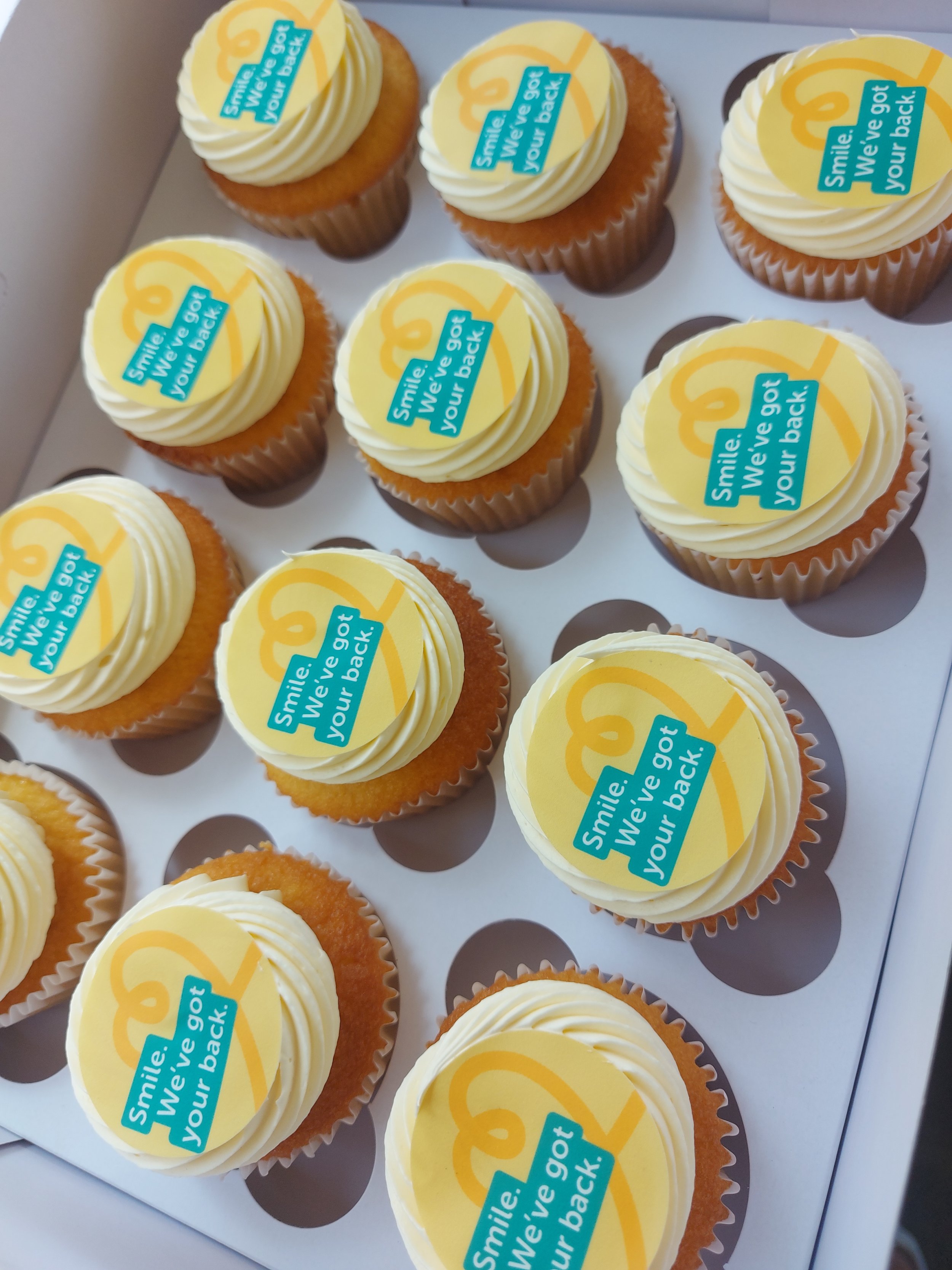 A box of cupcakes with yellow icing and a circular edible decoration that reads 'Smile. We've got your back!' in blue text on a yellow background.