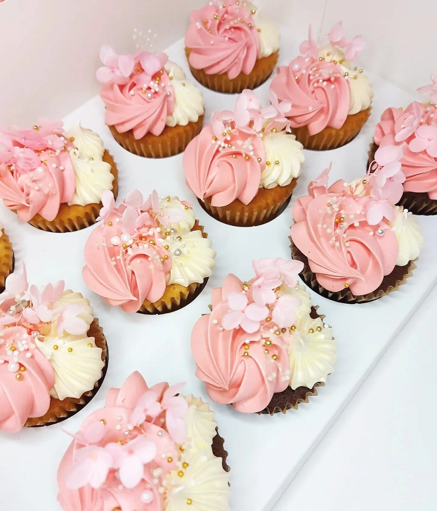 Assorted cupcakes decorated with pink and white frosting, edible pink flower decorations, and gold and pearl sprinkles.