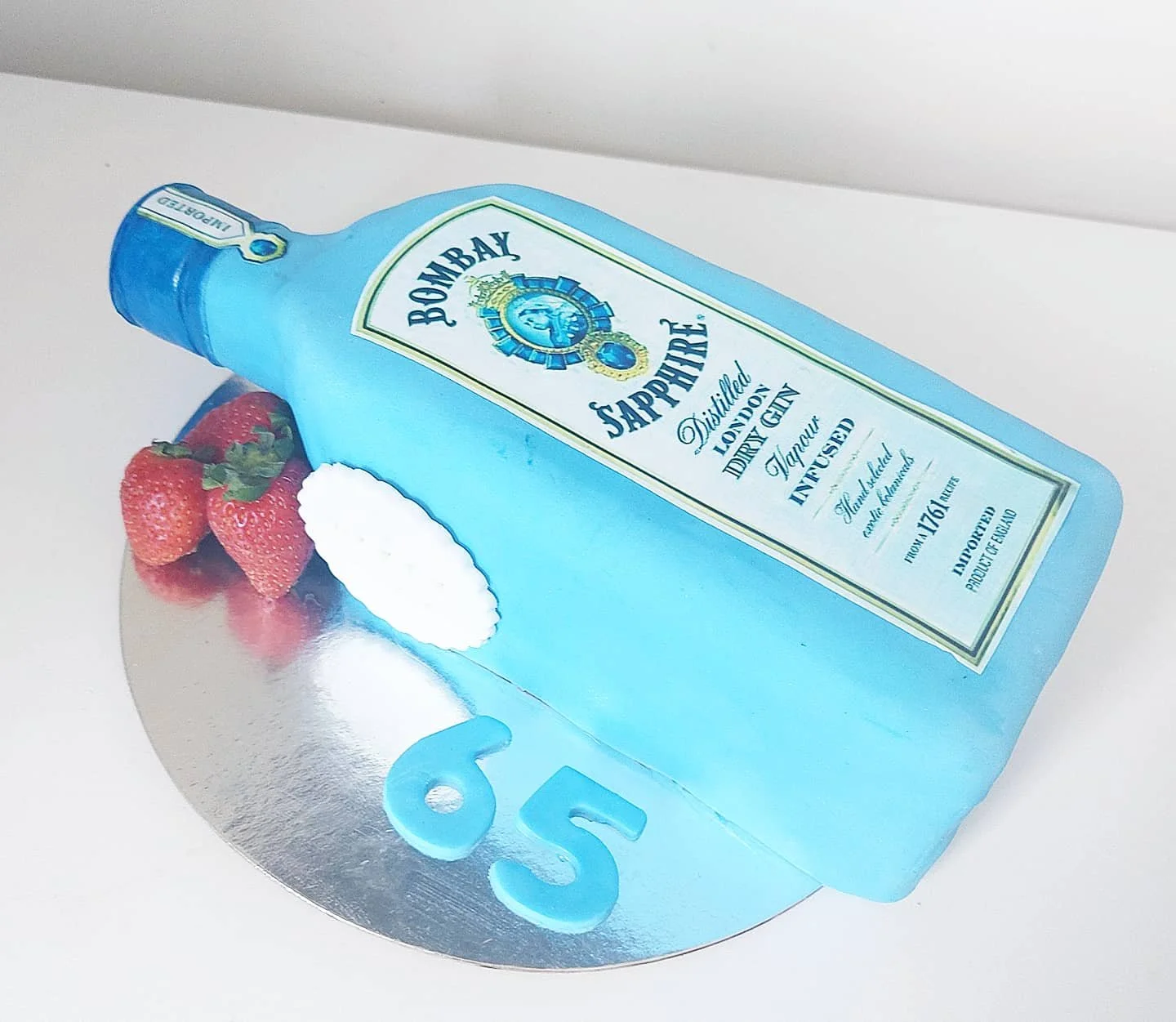 A shaped cake designed to look like Bombay Sapphire gin bottle, with strawberries and white cream decoration, placed on a round silver cake board with the number 65 on it.