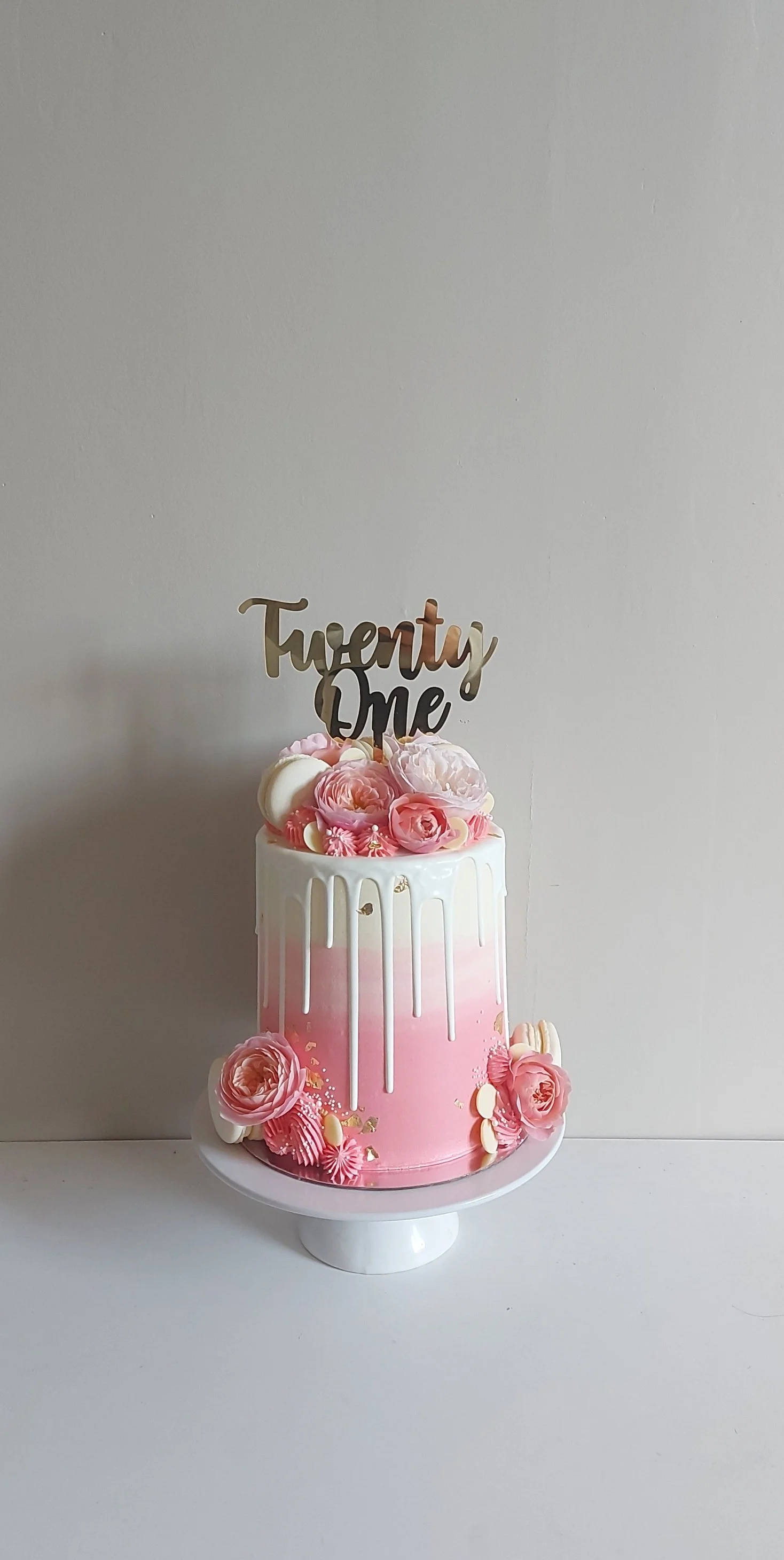 Pink birthday cake with white drip icing, decorated with pink and white flowers, gold accents, and topped with a "Twenty One" cake topper.