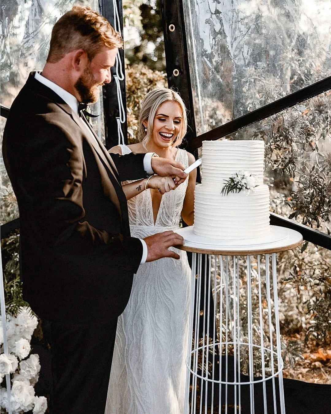 A newlywed couple in formal attire cutting a wedding cake together. The bride wears a white dress, and the groom wears a black tuxedo. The cake is white with textured frosting and small floral decorations, placed on a round wooden table with white me
