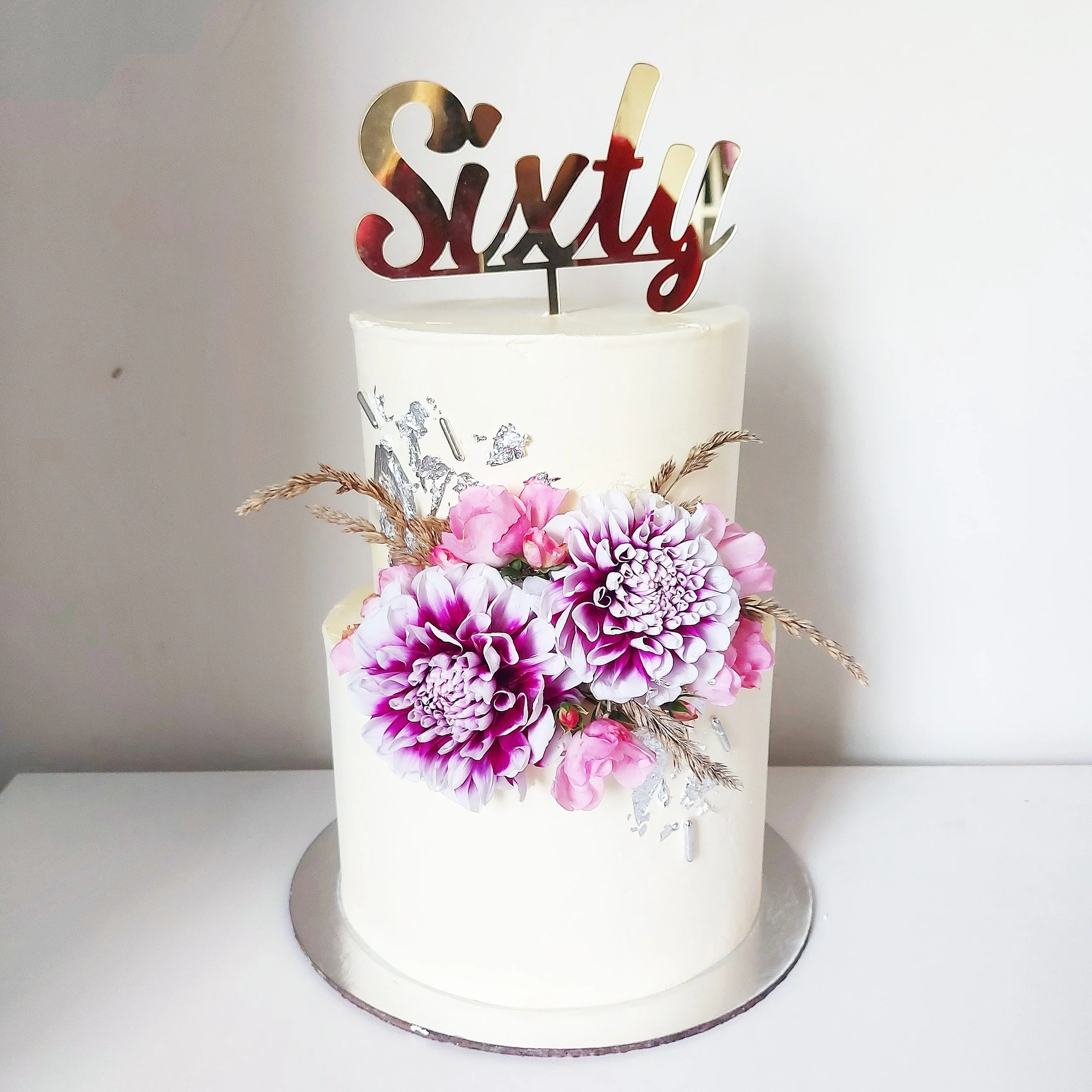A white birthday cake with pink and purple flowers and a gold topper that reads 'sixty'