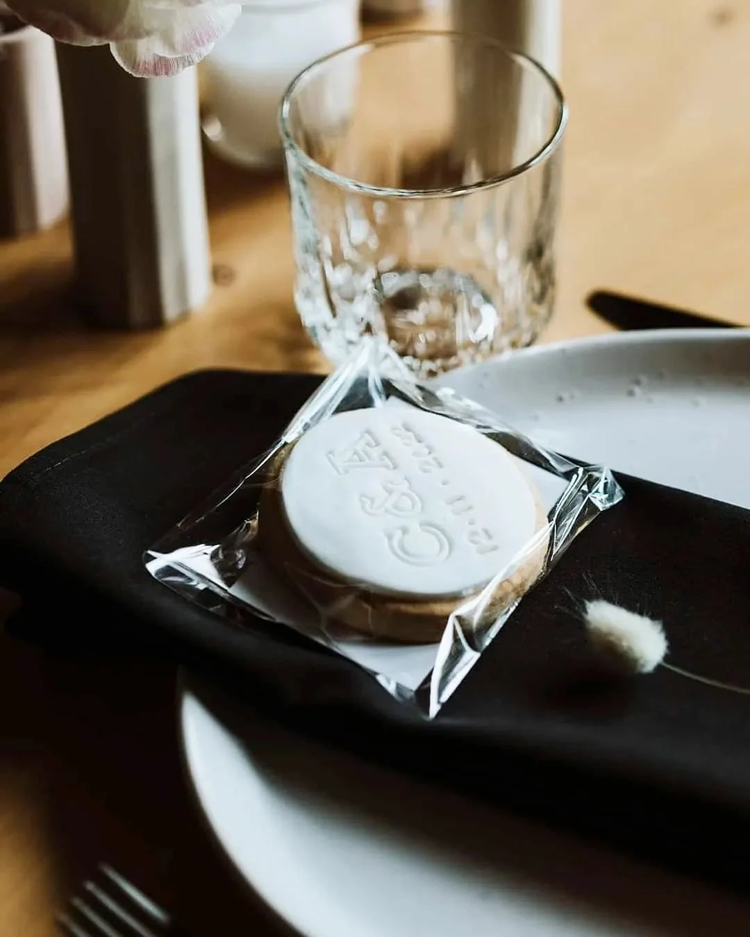 Close-up of a packaged cookie on a black napkin, glass of water, and white plate on a wooden table.