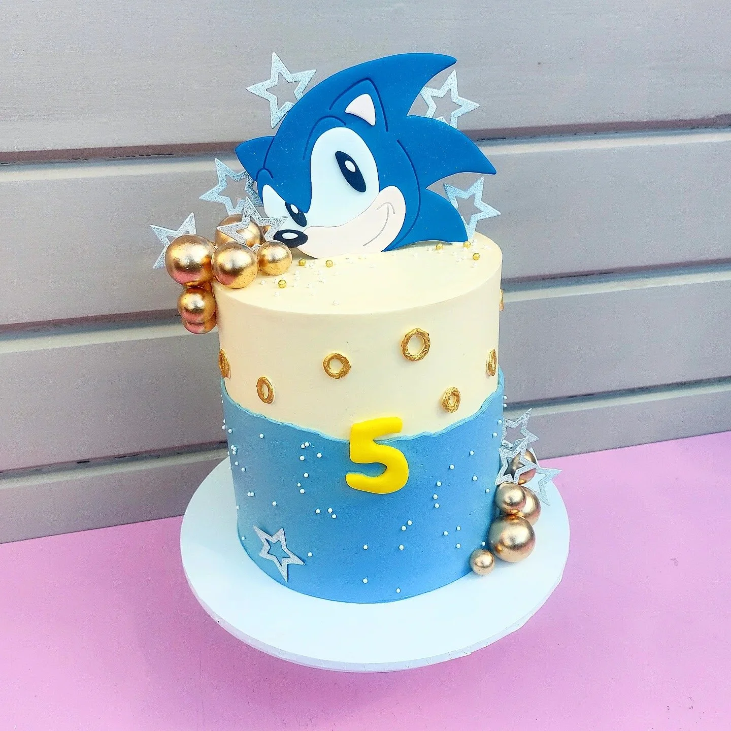 Birthday cake decorated in Sonic the Hedgehog theme with blue and cream frosting, gold and silver balls, star-shaped decorations, and a Sonic topper with the number 5.