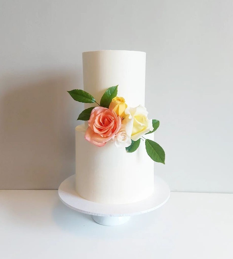 A white, two-tiered cake decorated with fresh pink, yellow, and white roses and green leaves.
