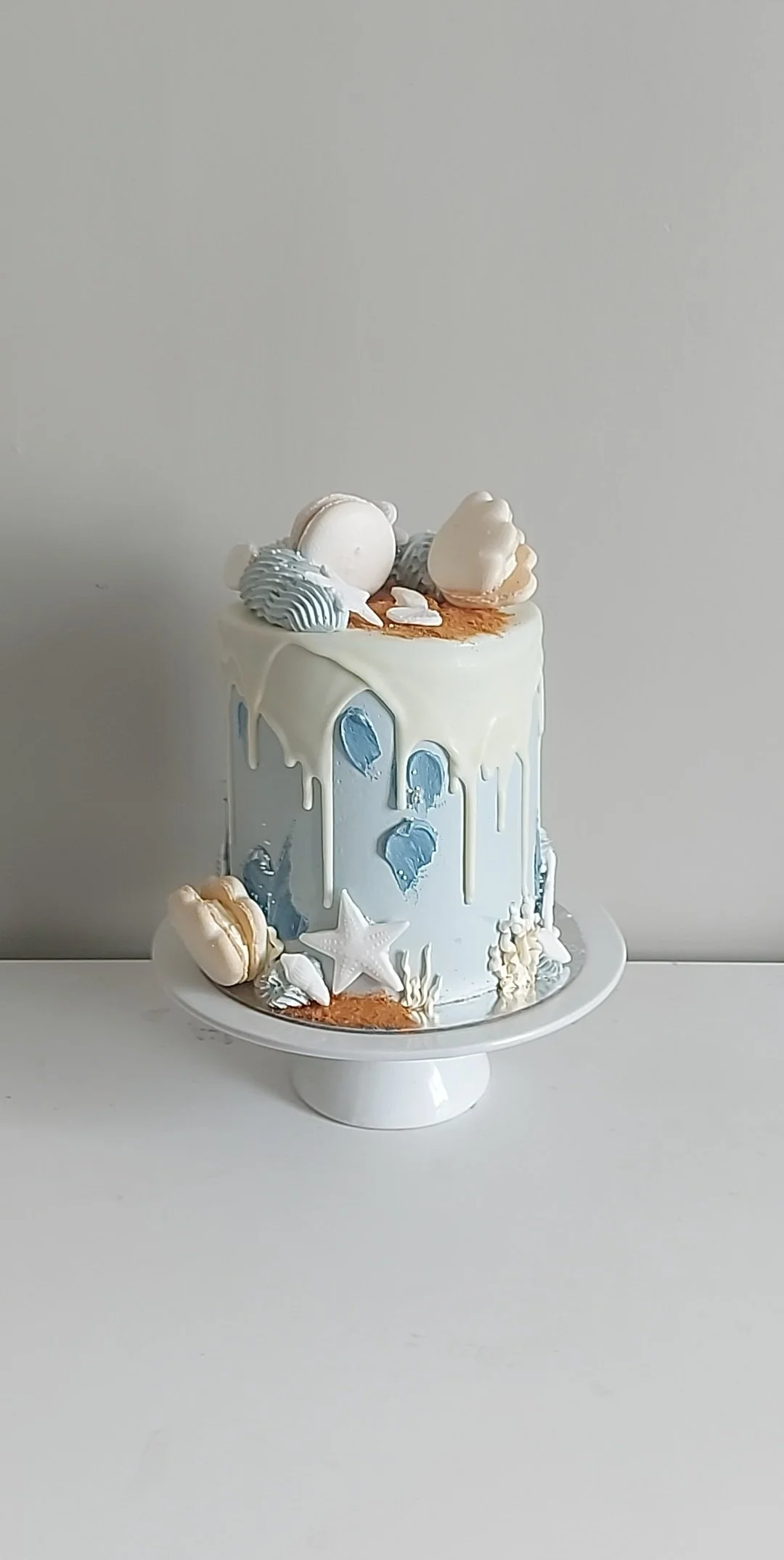 Decorative white cake with blue and white icing, featuring seashell, starfish, and sandy accents on a white cake stand.