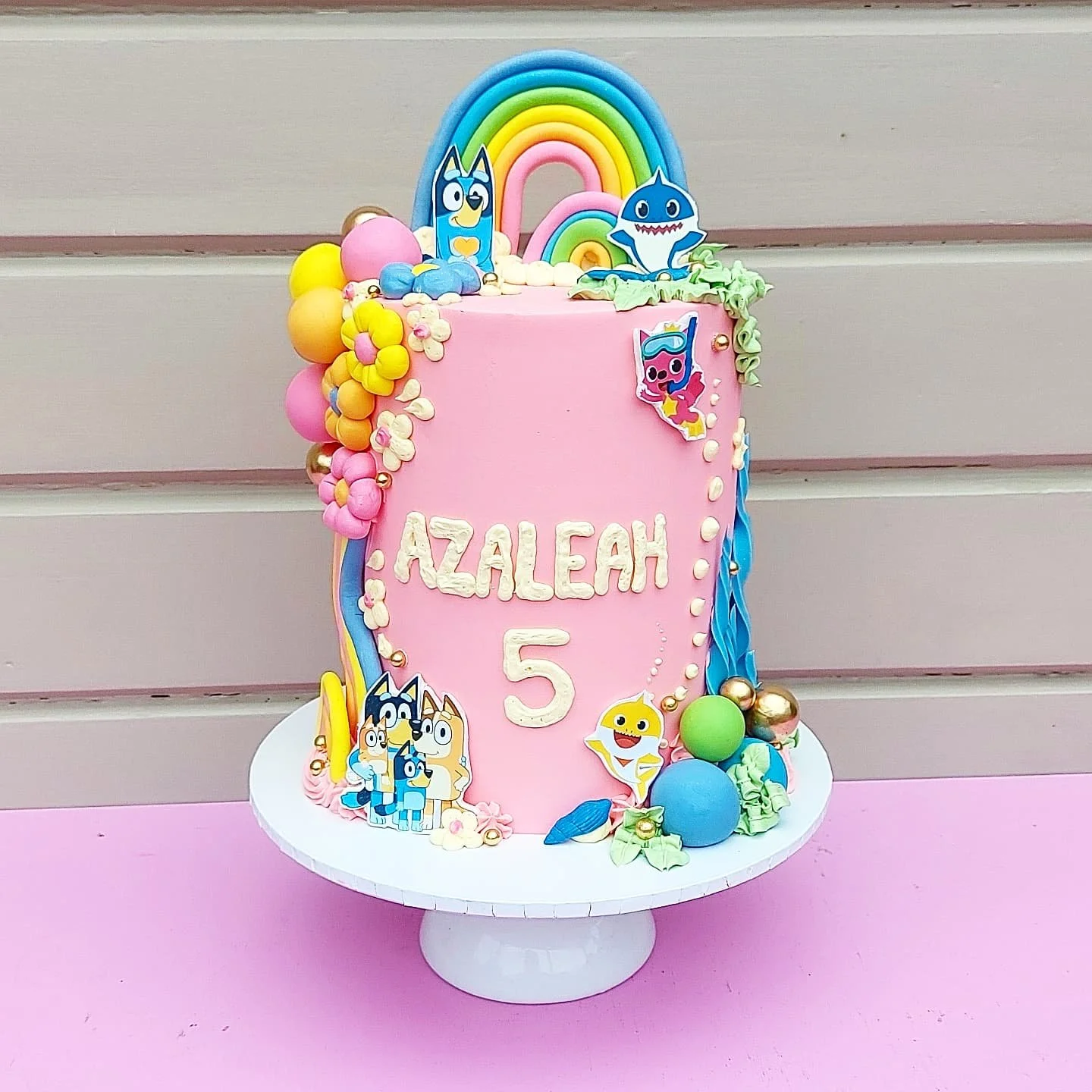 Pink birthday cake with colorful decorations, including rainbows, cartoon characters, flowers, and balls. The cake has the name "Azaleah" and the number "5" on it, placed on a white cake stand against a pink surface and beige wall background.