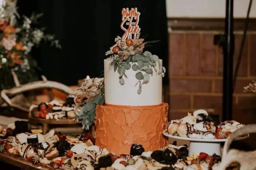 A two-tiered wedding cake with a white top tier decorated with leaves and a rust-colored bottom tier with textured frosting, topped with a cake topper that reads "Mr. & Mrs. Sull". The cake is surrounded by various desserts and strawberries.