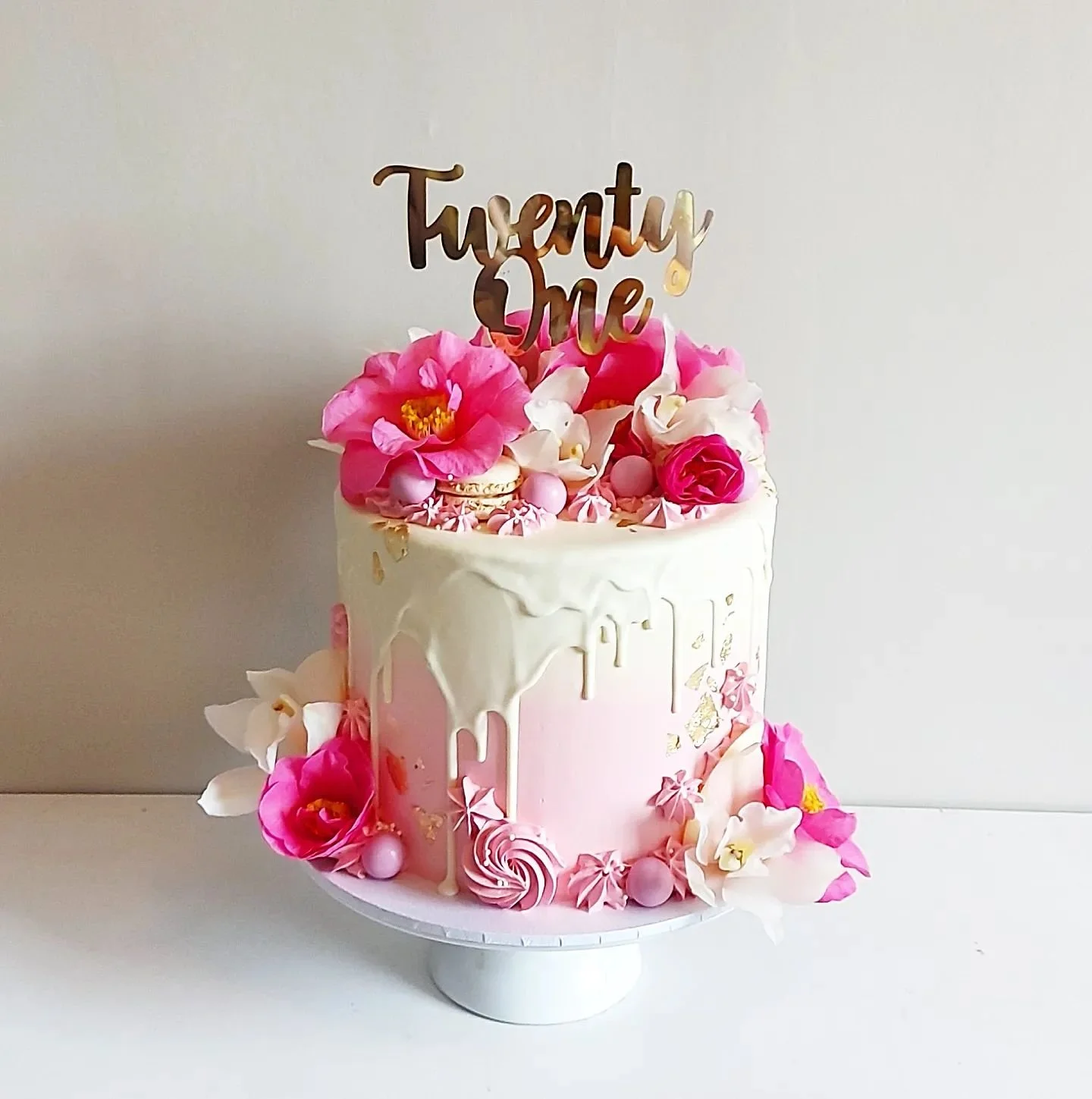 Pink and white birthday cake decorated with flowers, macarons, and a golden "Twenty One" cake topper.