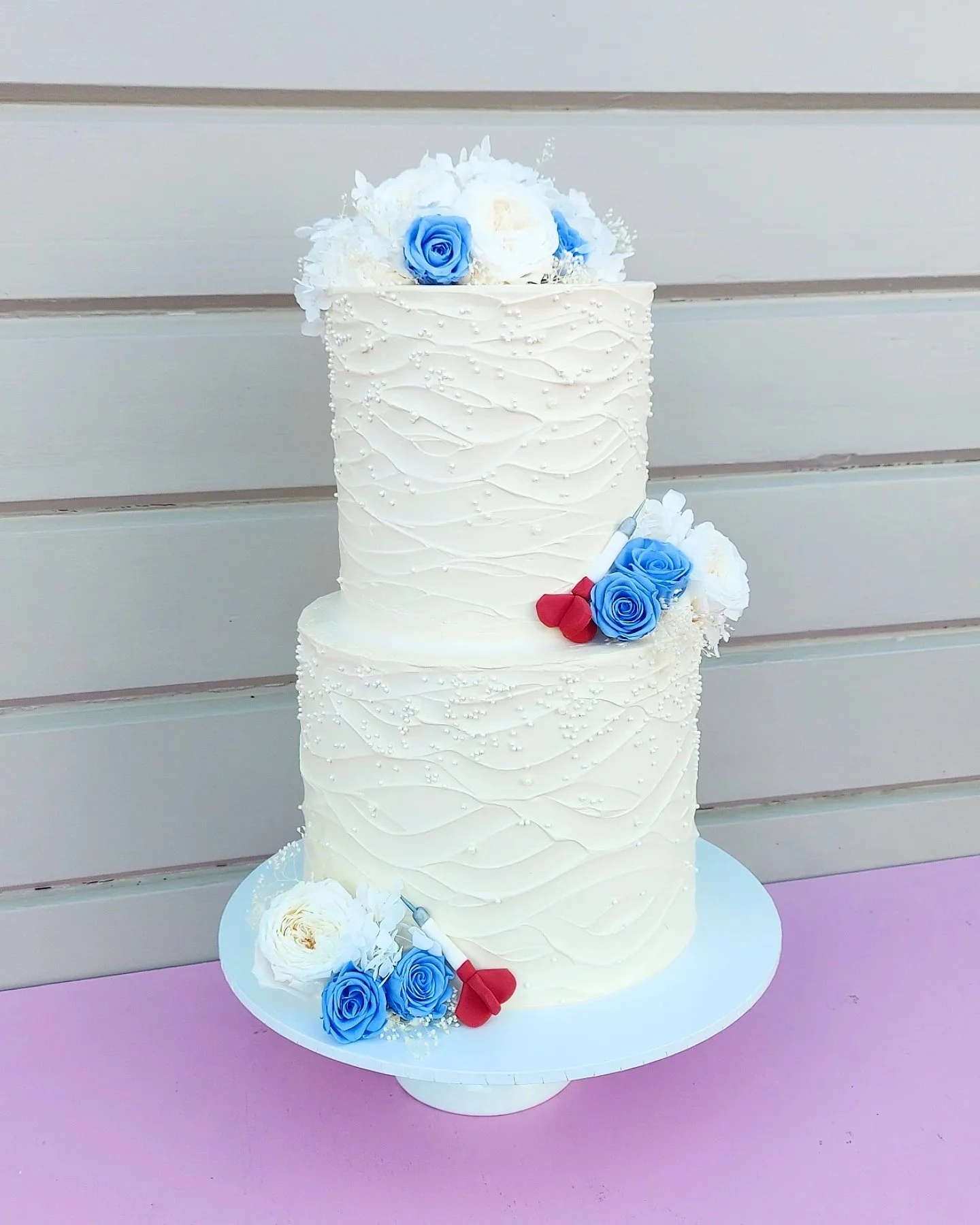 A 2-tier white wedding cake decorated with blue and white roses, white hydrangeas, and small pearl-like accents, placed on a pink surface against a gray wooden background.