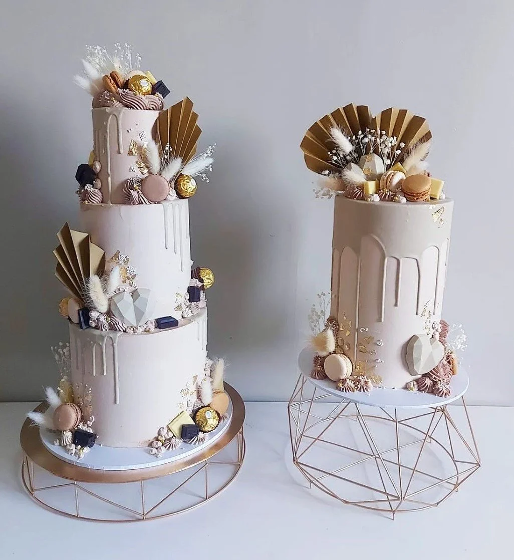 Two elegant, cream-colored cakes decorated with gold, pink, and white accents. The larger, tiered cake on the left is topped with assorted chocolates, macarons, and decorative elements. The smaller cake on the right features similar decorations. Both