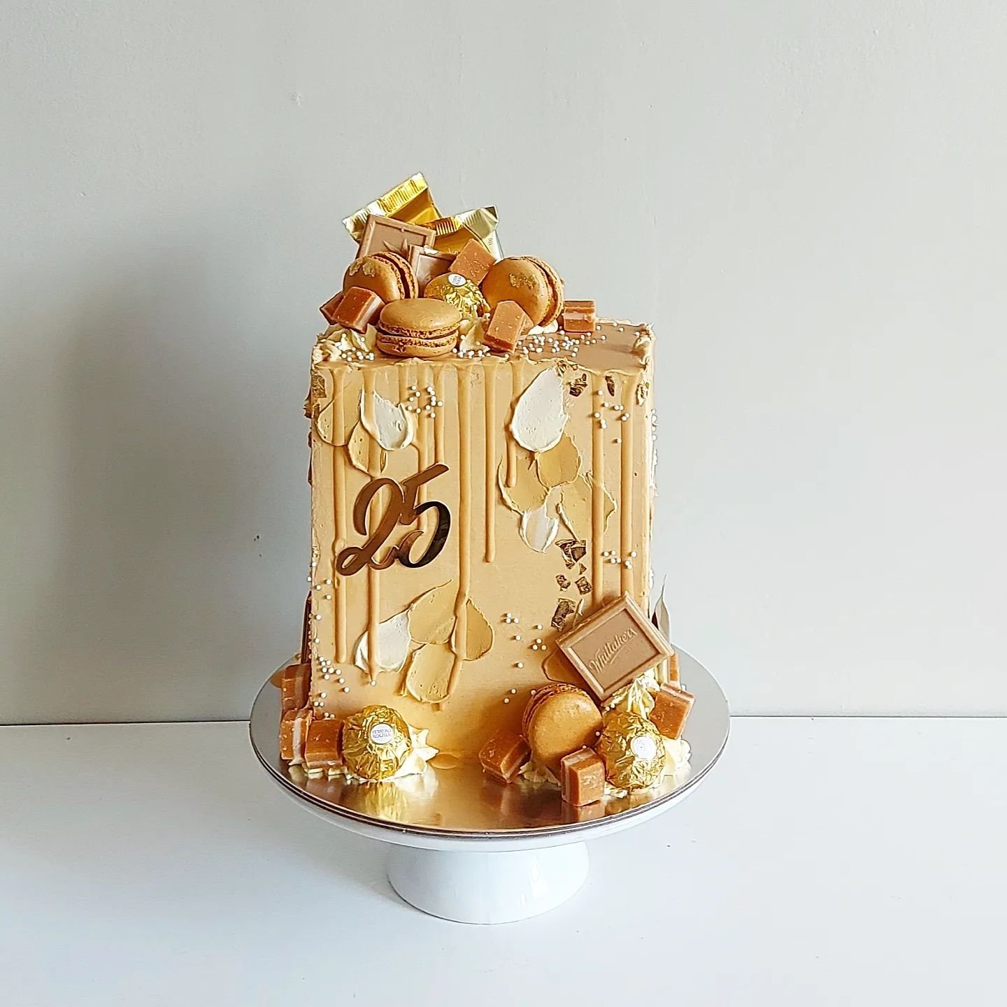 Elegant beige birthday cake with gold and chocolate decorations, topped with macarons, chocolates, and a '25' topper on a white cake stand.