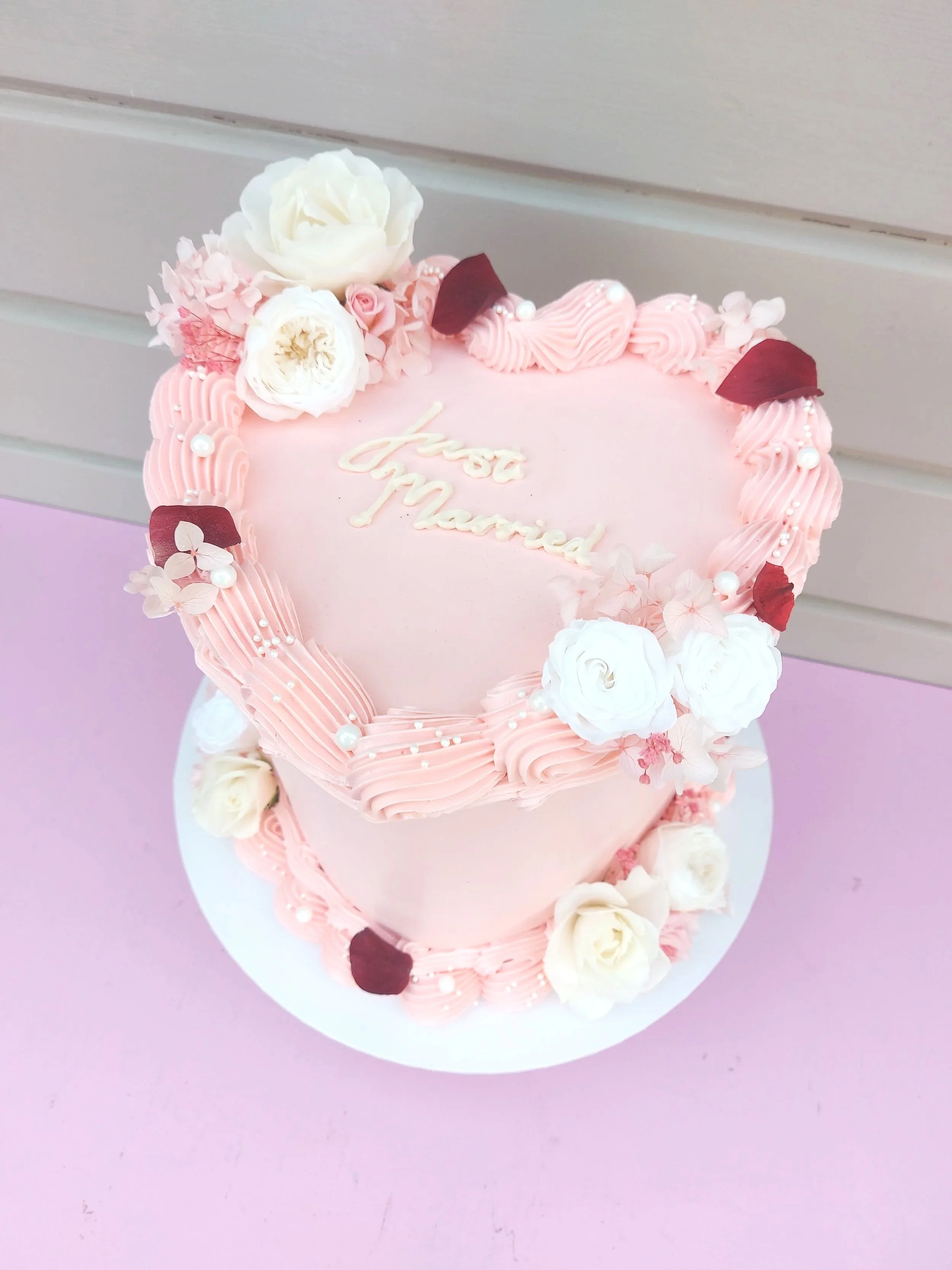A pink heart-shaped cake decorated with white and pink flowers, icing pearls, and a message that says 'just married' in white icing.