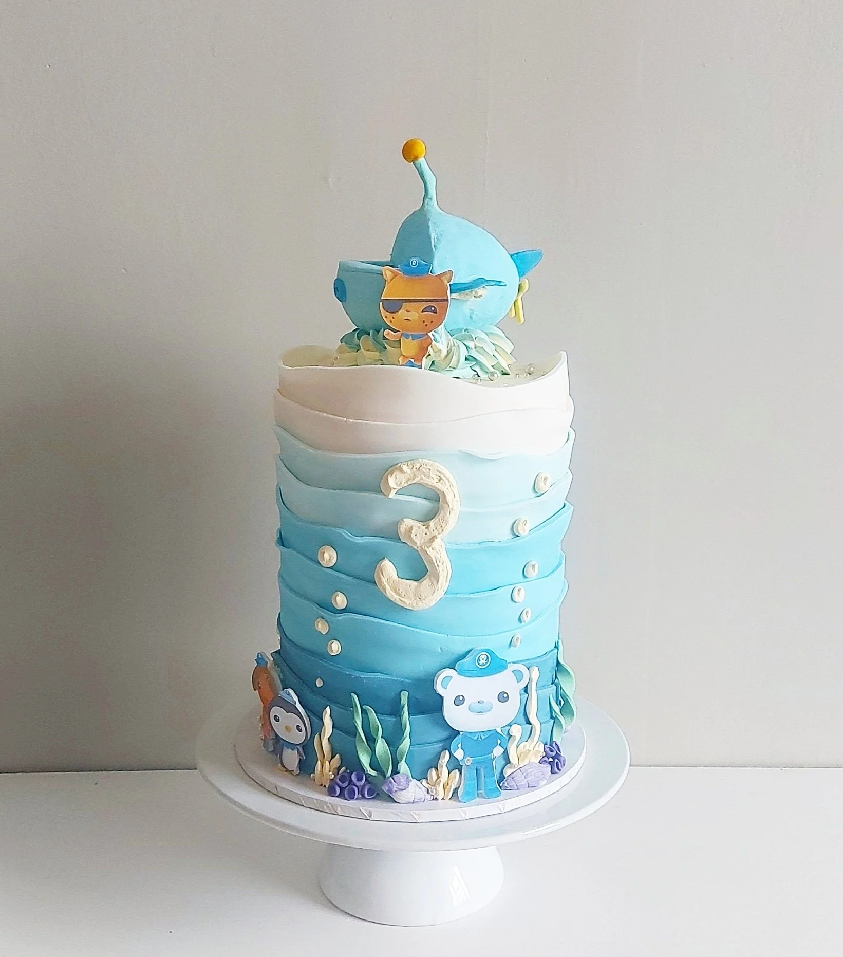 A multi-tiered cake decorated with an ocean theme, including sea creatures, seaweed, and a nautical boy figurine, with a prominent white number three on the front, against a plain background.