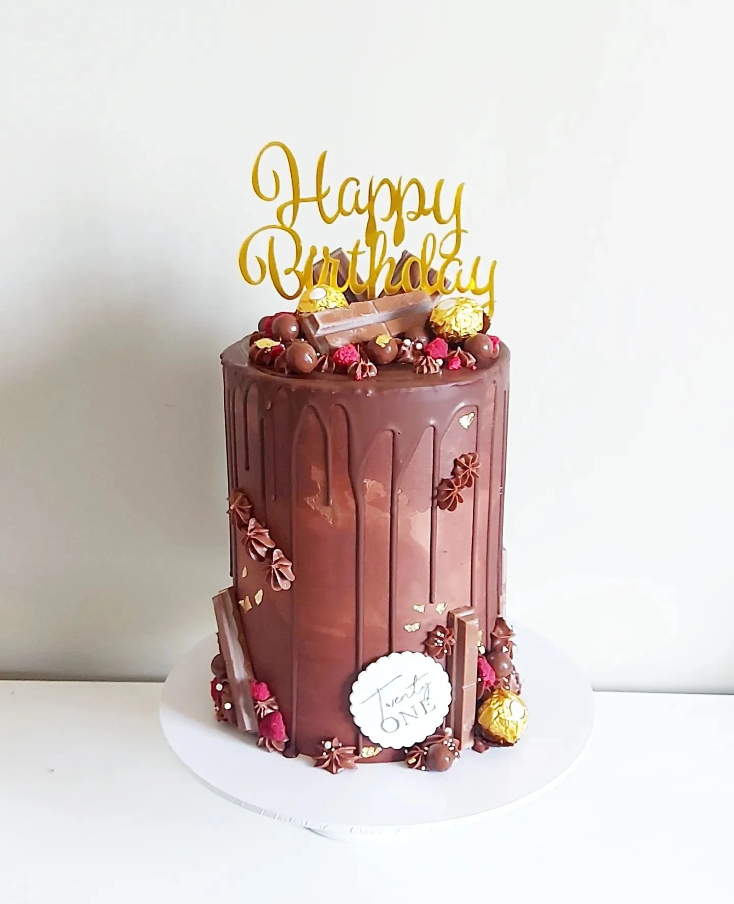 Chocolate birthday cake with a gold "Happy Birthday" topper, decorated with chocolate balls, chocolate bars, pink and red candies, and gold accents. The cake has a label reading "Twenty ONE."