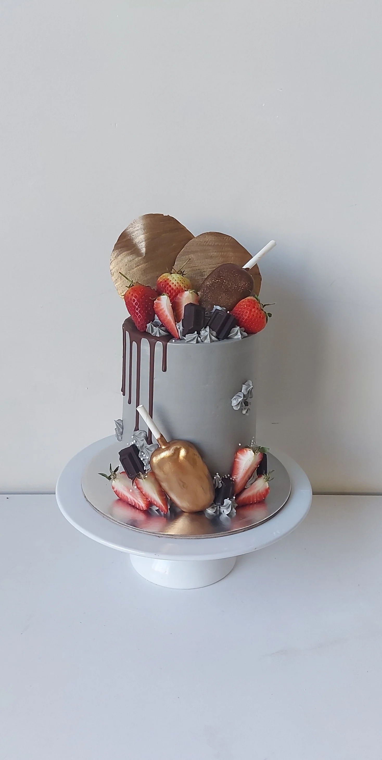 Two-tiered cake decorated with strawberries, chocolate pieces, gold-covered fruit, and gold hearts, with a white and silver color scheme.