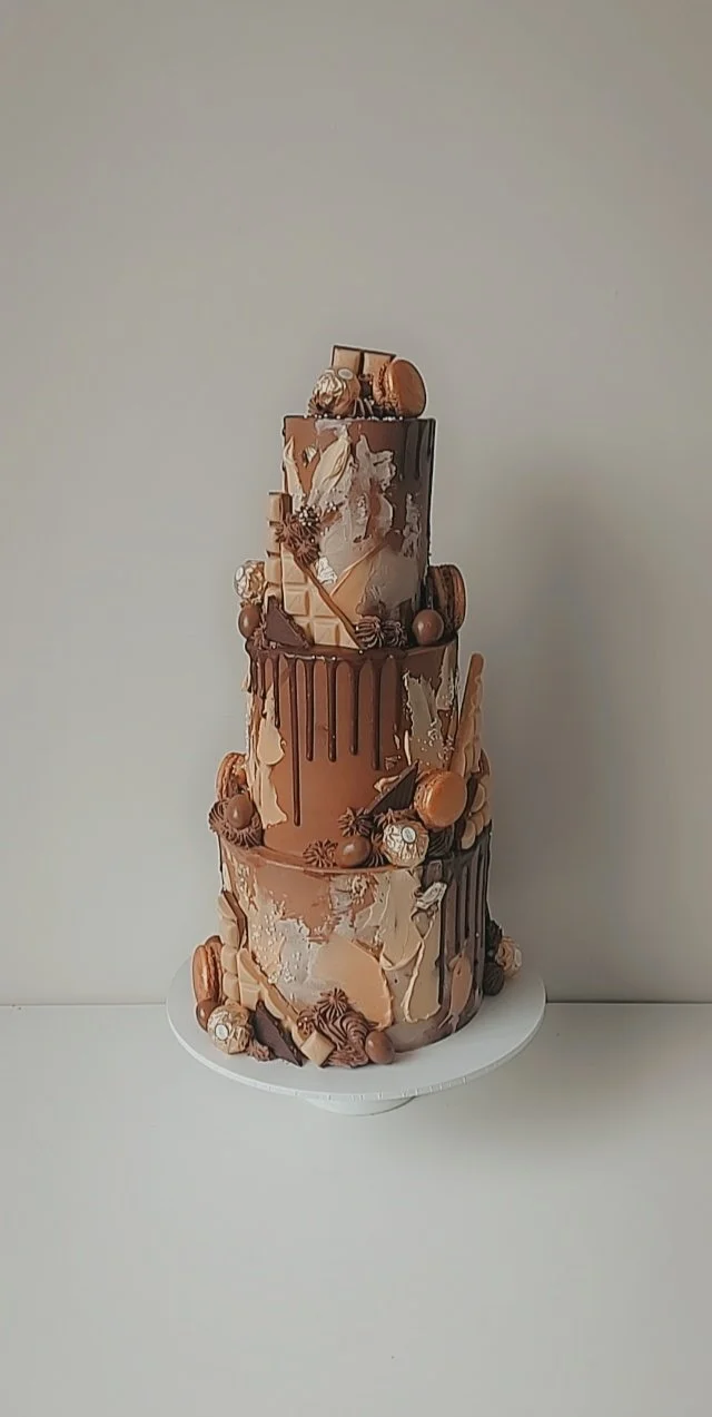 Three-tiered chocolate cake decorated with chocolate drip, macarons, chocolate pieces, and decorative chocolate shavings on a white cake stand against a plain background.