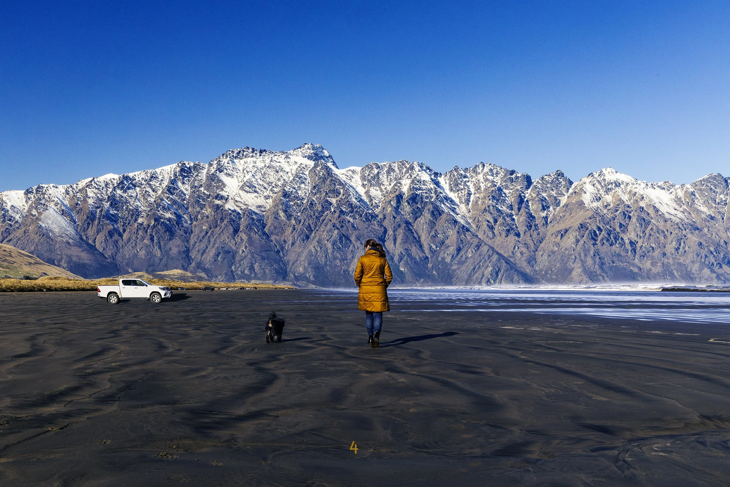 A woman and dog walk on a beach in front of The Remarkables.
