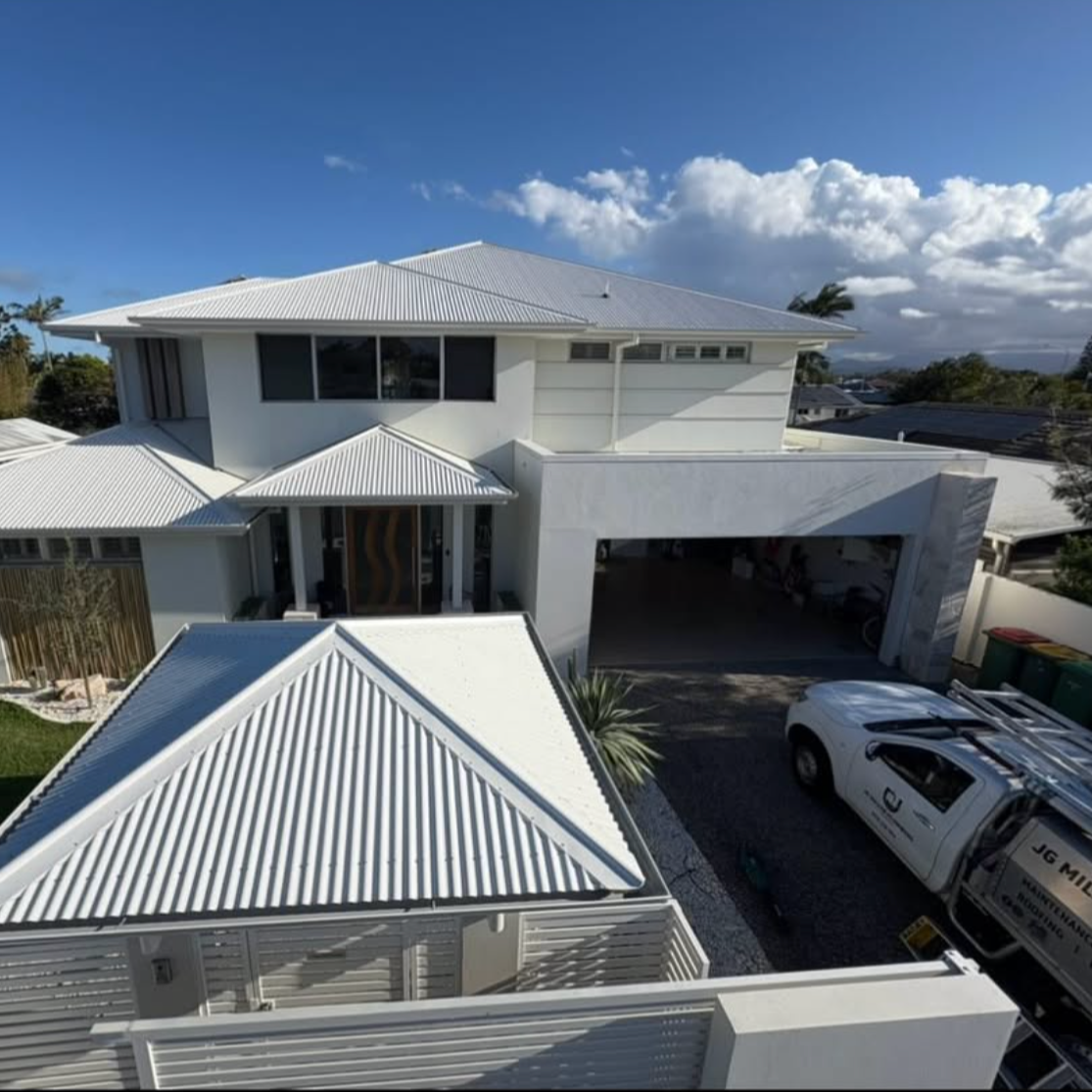 White modern two-story house with metal roof, large windows, and an open garage area. A white truck is parked outside, and there are green trash bins and plants nearby. Clear blue sky with some clouds in the background.