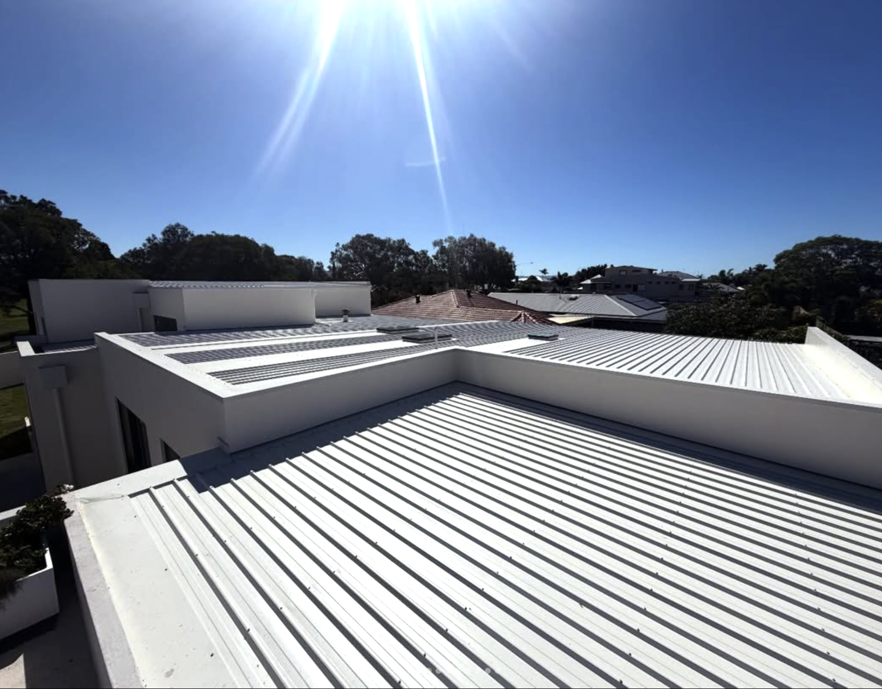 View of modern white buildings with metal roofs under a bright sun in a residential area with trees and neighboring houses.