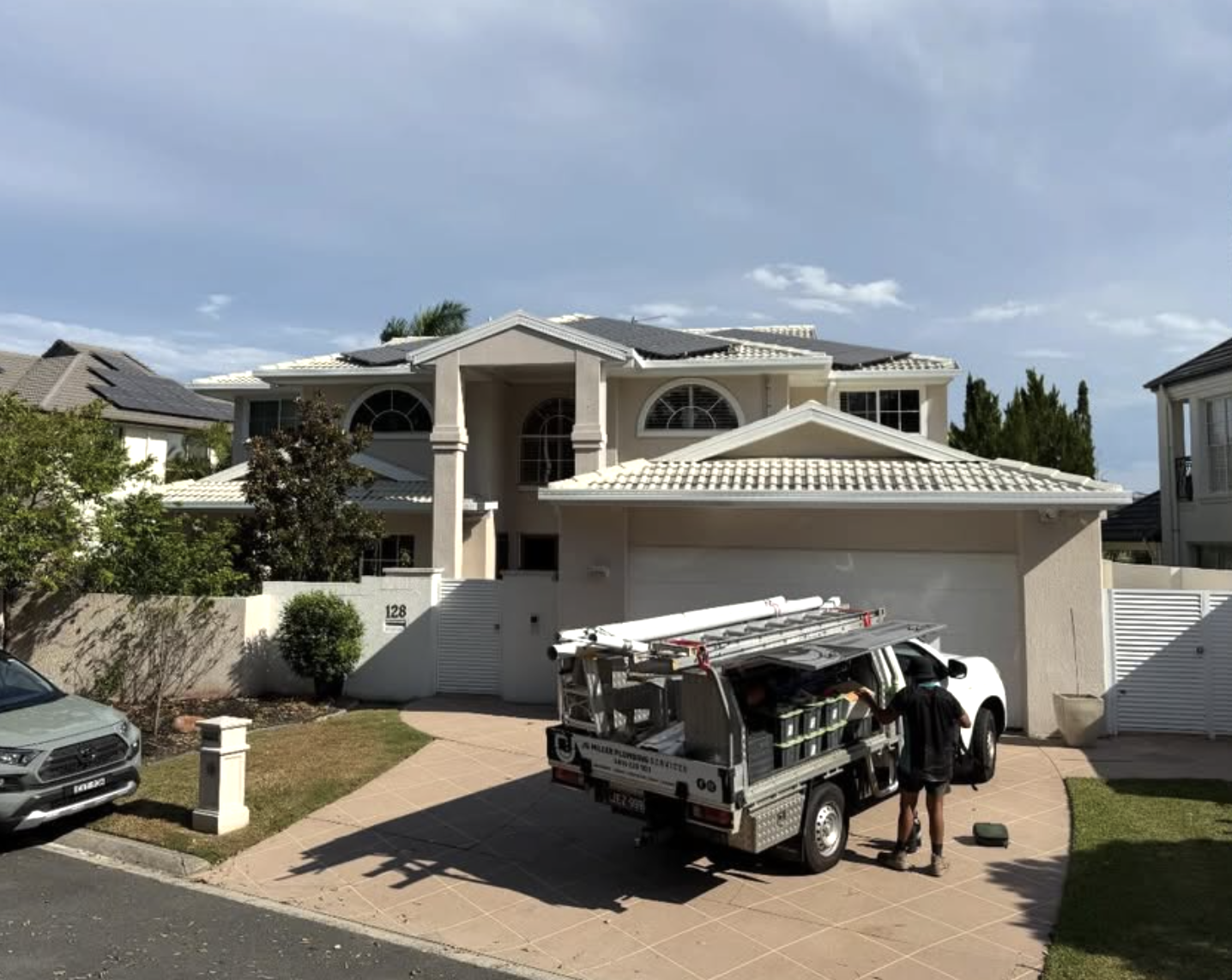 A residential house with a driveway, a worker standing next to a service truck, and a car parked on the street.