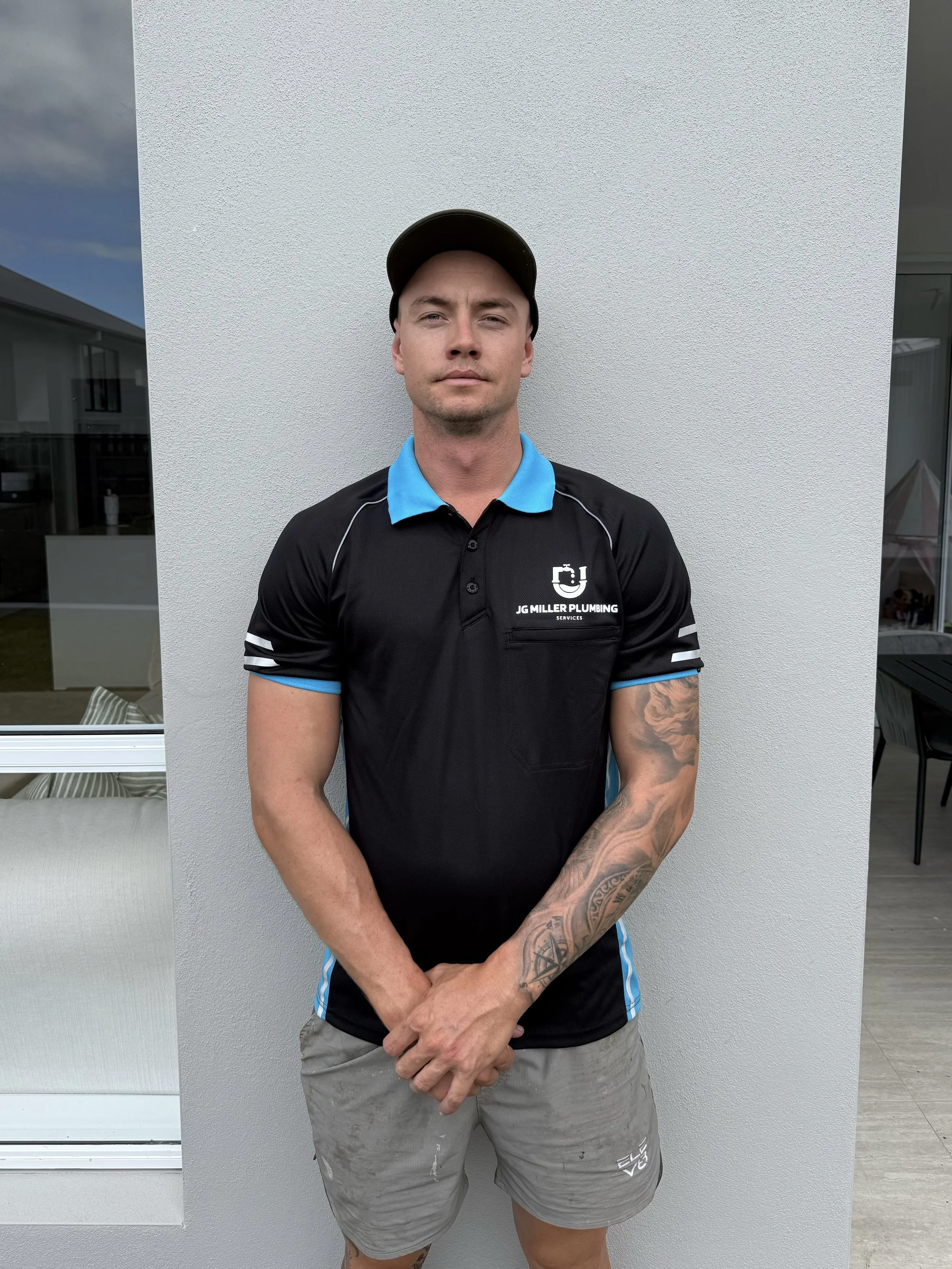 A young man in a black polo shirt with blue accents and a logo that says "JG MILLER PLUMBING SERVICES" stands outdoors against a white wall, wearing a black cap and gray shorts, with tattooed arms, looking at the camera.