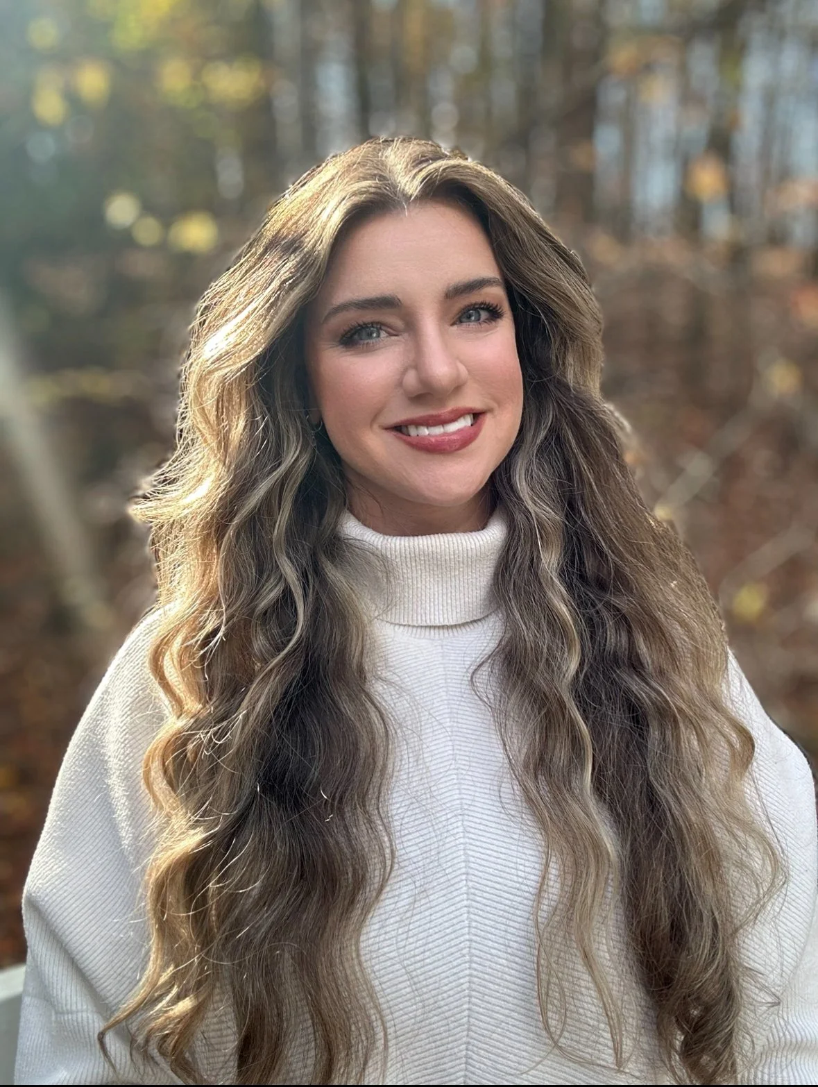A woman with long wavy light brown hair and blue eyes smiling outdoors in a forest during autumn, wearing a white turtleneck sweater.