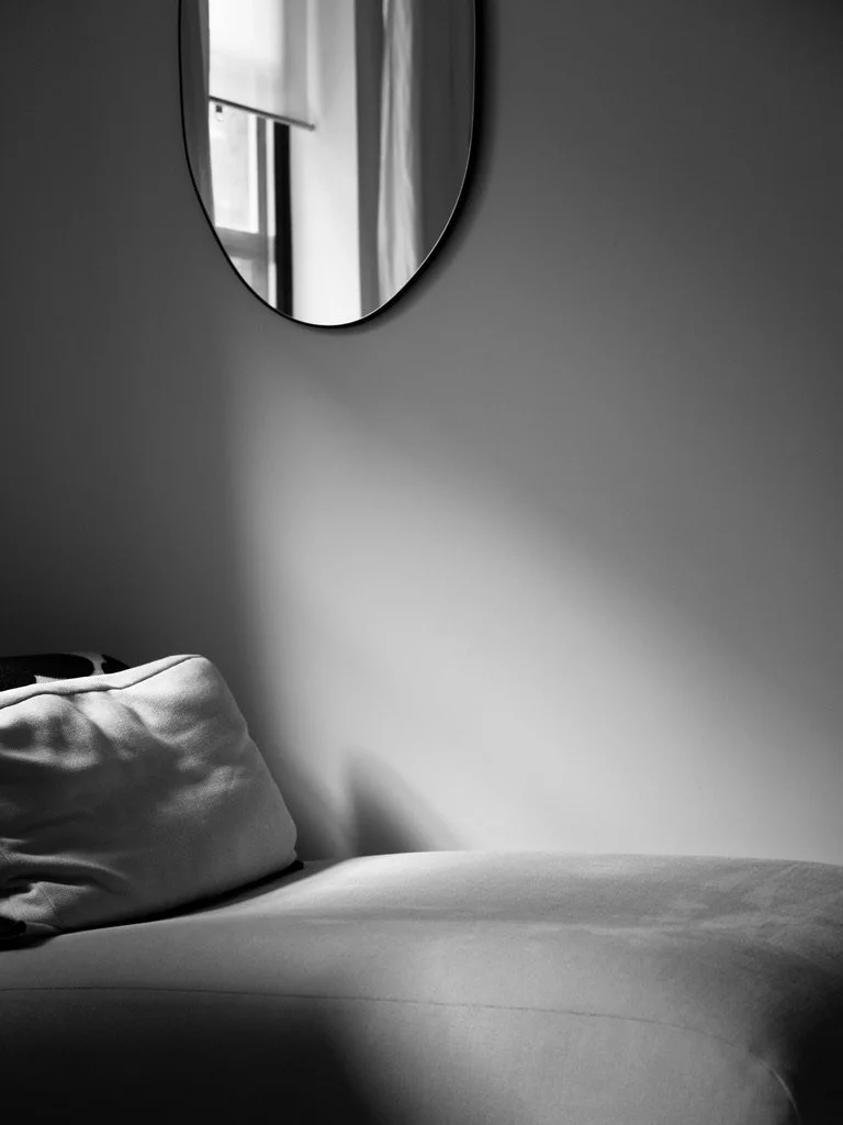 A black and white photo of a bed with a pillow on it, a wall mirror, and a window with curtains reflecting in the mirror.