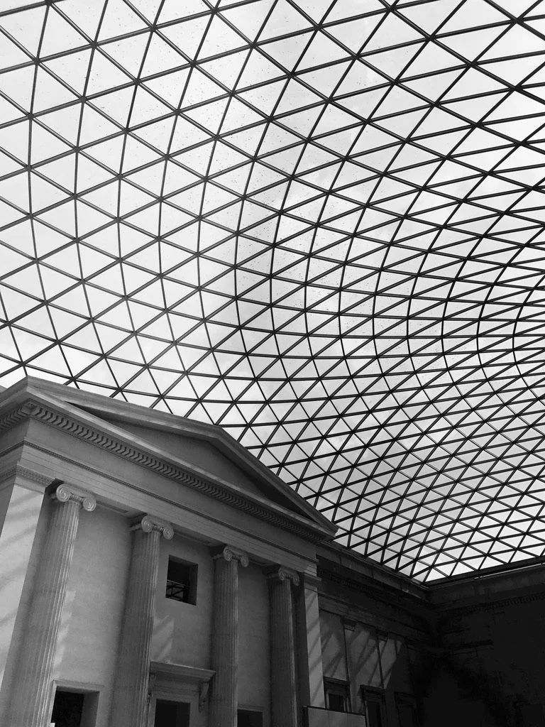 The image shows the interior of a building with classical architecture featuring tall columns and a triangular pediment, under a modern glass and steel dome ceiling with a grid pattern.