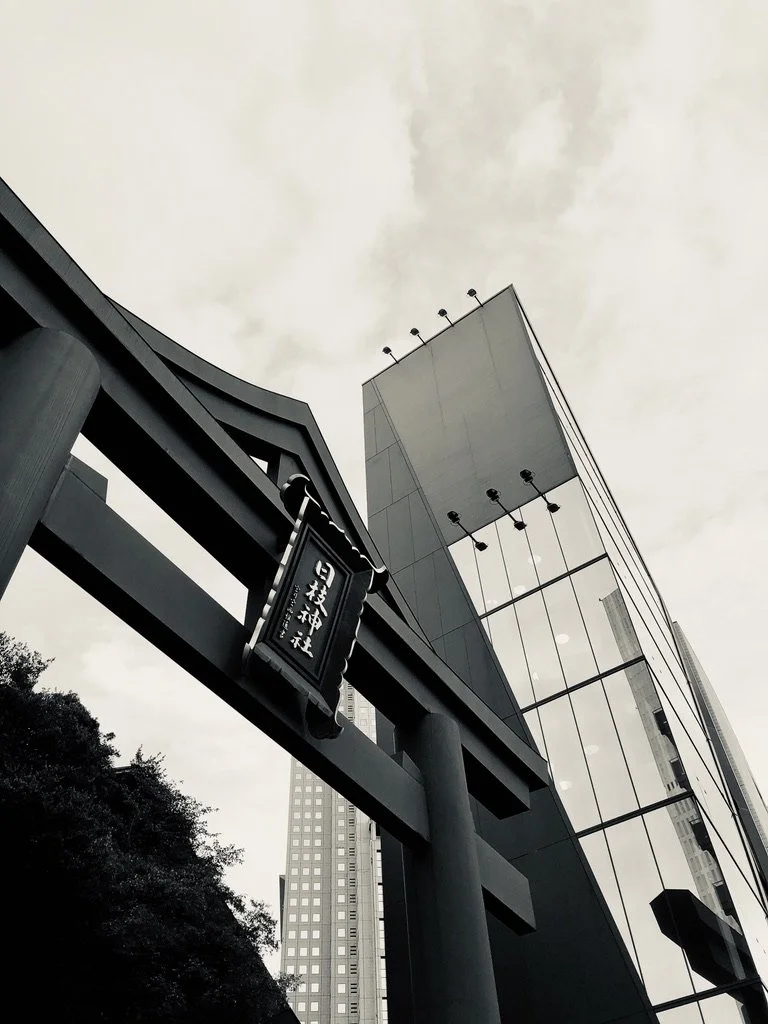 Low-angle view of a modern building with glass windows and a torii gate with Japanese characters in an urban setting, cloudy sky in the background.
