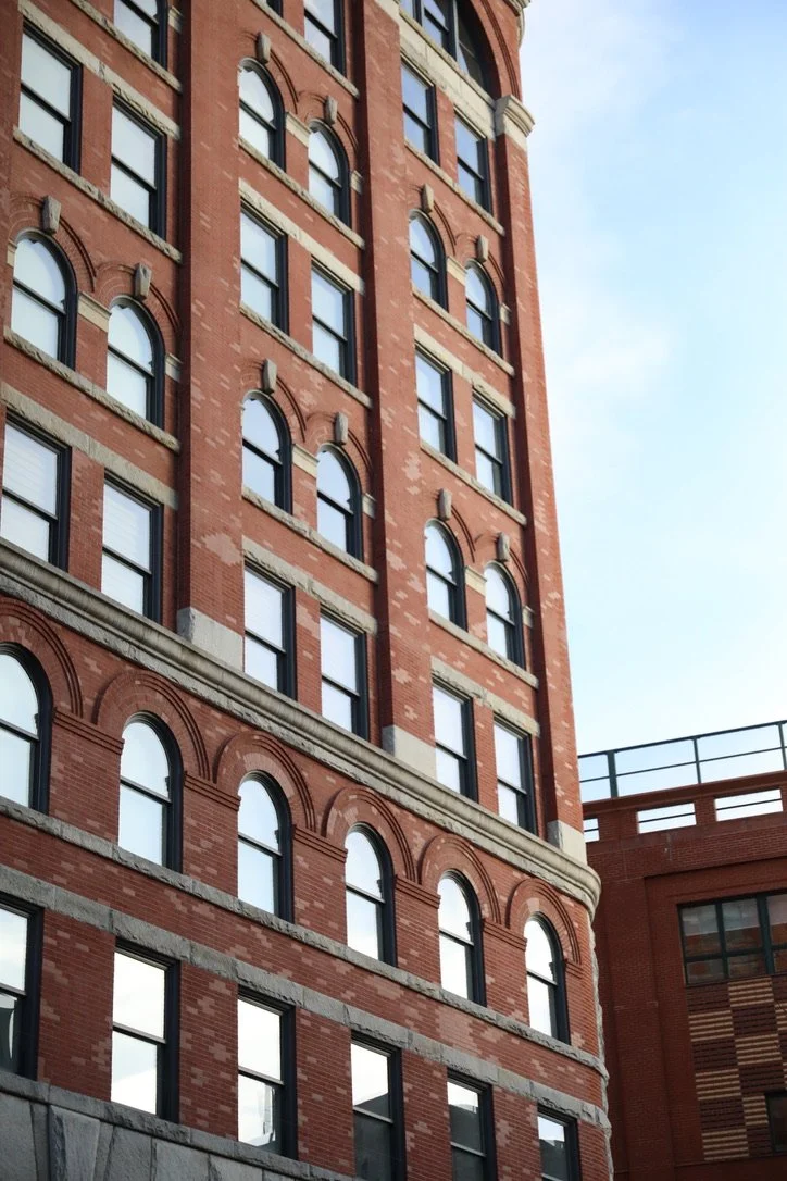 A tall, red brick building in New York City with multiple arched windows, some with black frames and some with white blinds, under a partly cloudy sky.