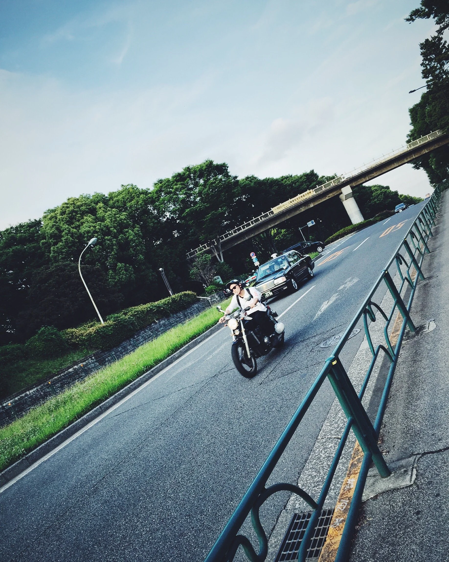 A person riding a motorcycle in Shibuya, Tokyo, Japan on a street, trees in the background, and an overpass overhead.