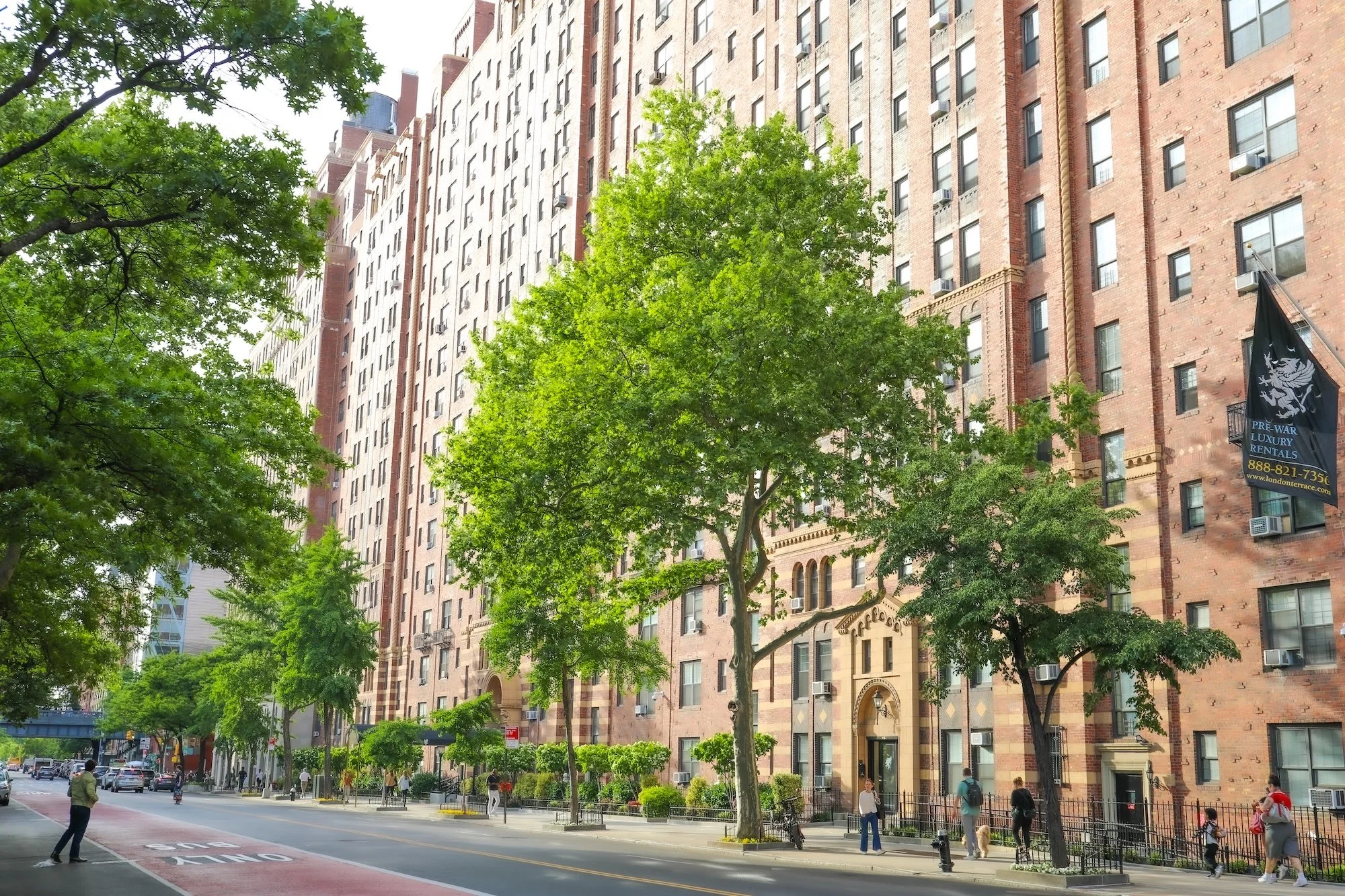 A city street scene with a large brick apartment building, trees along the sidewalk, and several pedestrians walking. A flag advertising luxury rentals is visible near the building.