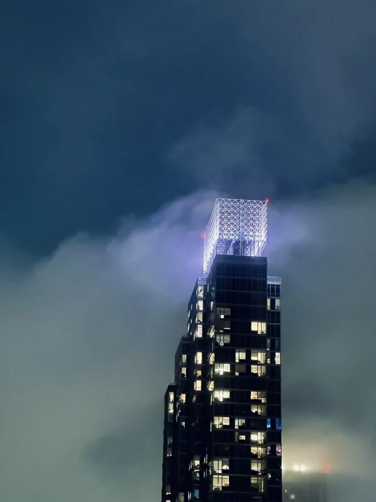 Nighttime view of a tall modern skyscraper with illuminated windows and a bright, illuminated crown at the top, shrouded in fog with some clouds in the background.