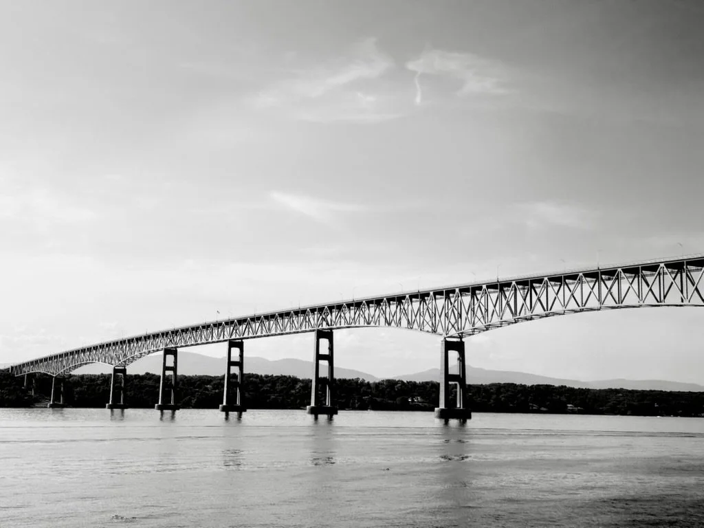 Black and white photo of a long bridge extending over a body of water, with mountains in the background and a cloudy sky overhead.