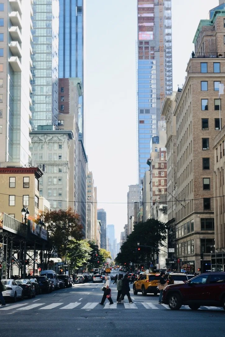 City street with tall buildings, pedestrians crossing, and cars parked along the sides.
