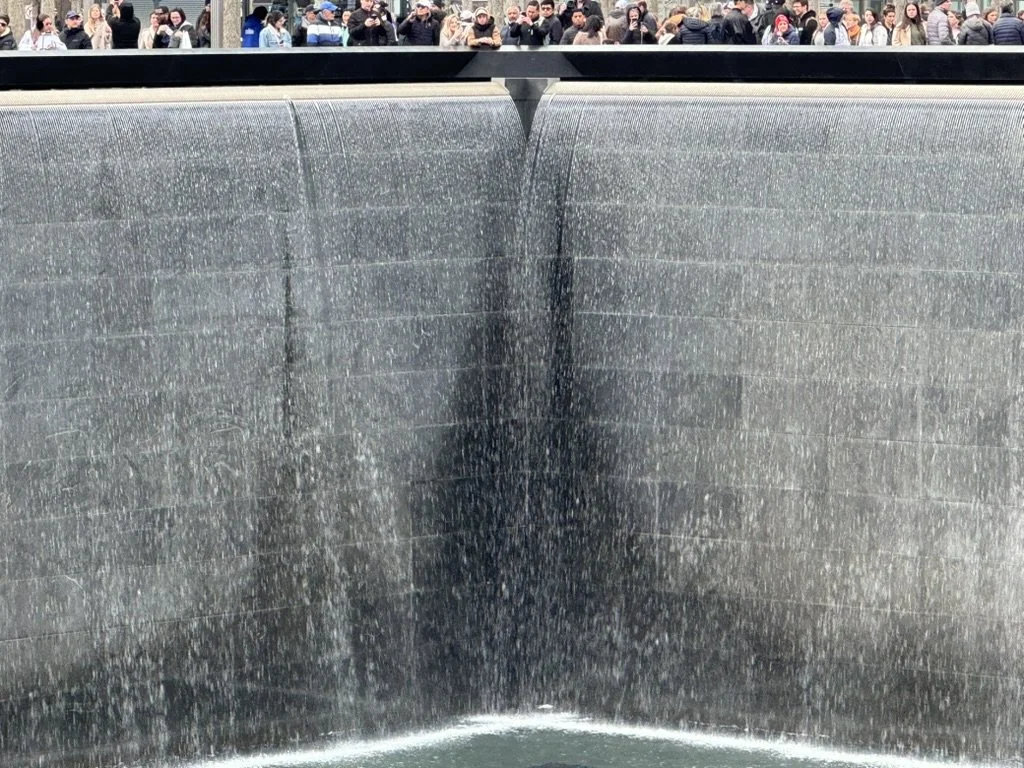 New York City 9/11 memorial wall with water cascading down its black surface and a crowd of people gathered in the background.