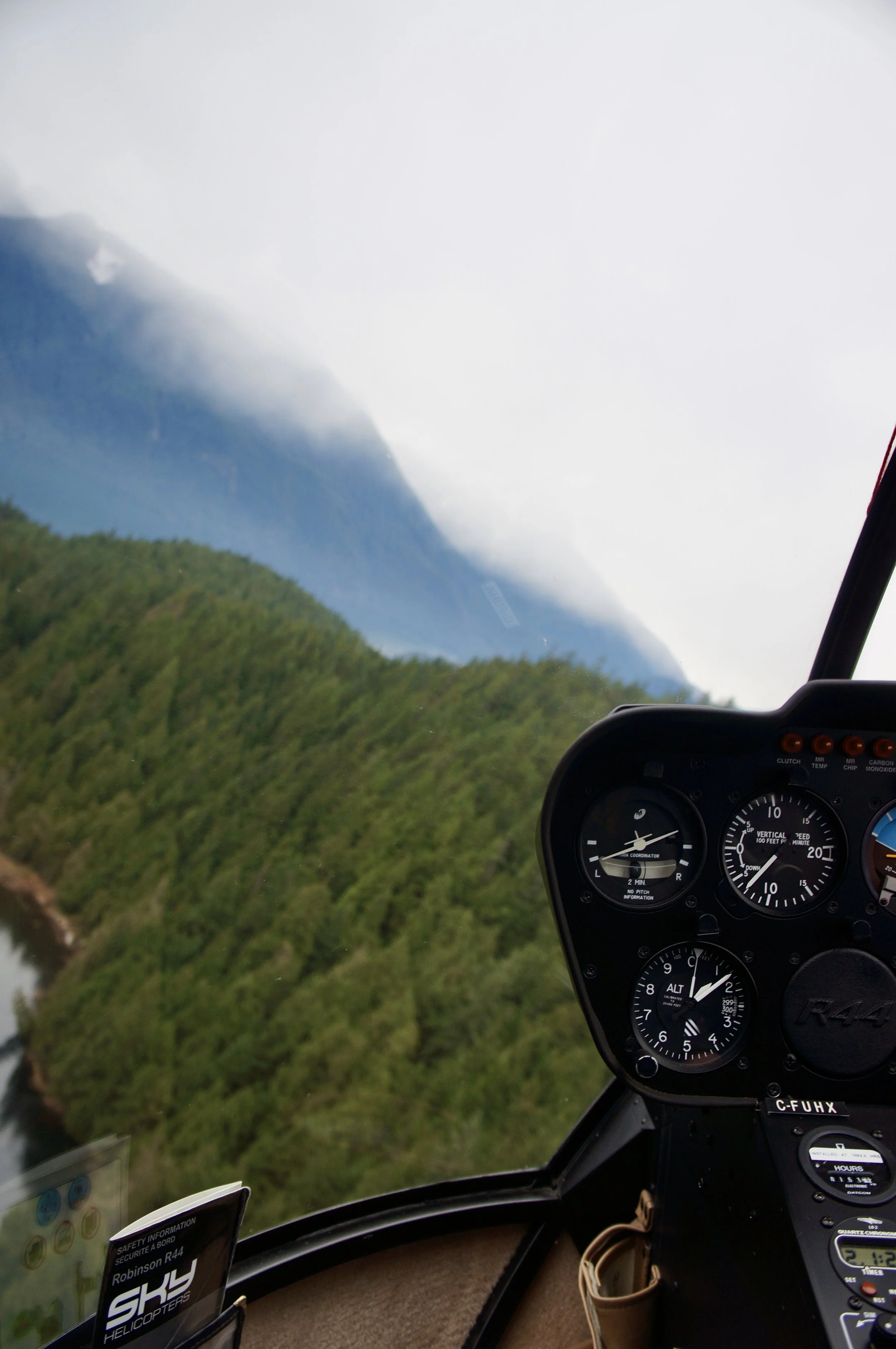 Inside the cockpit of a Robinson R44 helicopter, showing the instrument panel and partial view of the helicopter's dashboard with a scenic mountain landscape in the background.