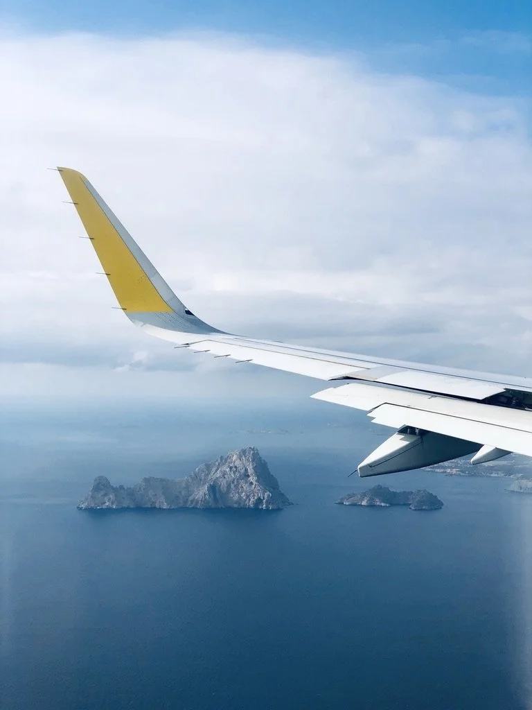 View from an airplane window showing the airplane wing and a large rocky island surrounded by water under a cloudy sky.