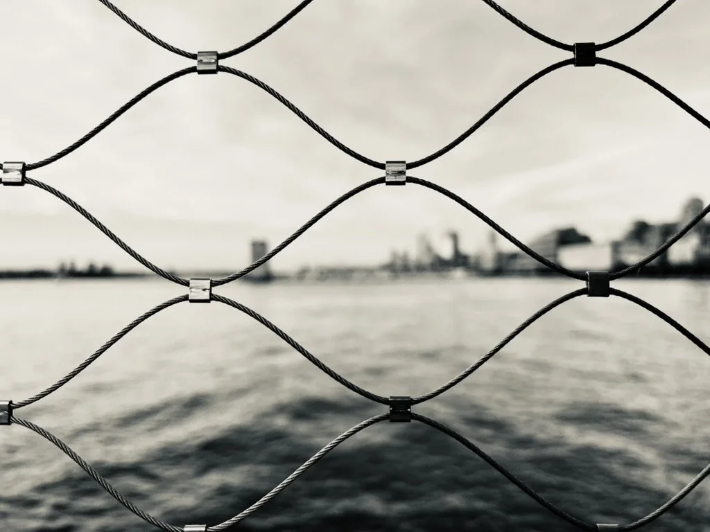 A chain-link fence with wire and metal connectors in front of a body of water and a city skyline in the background.