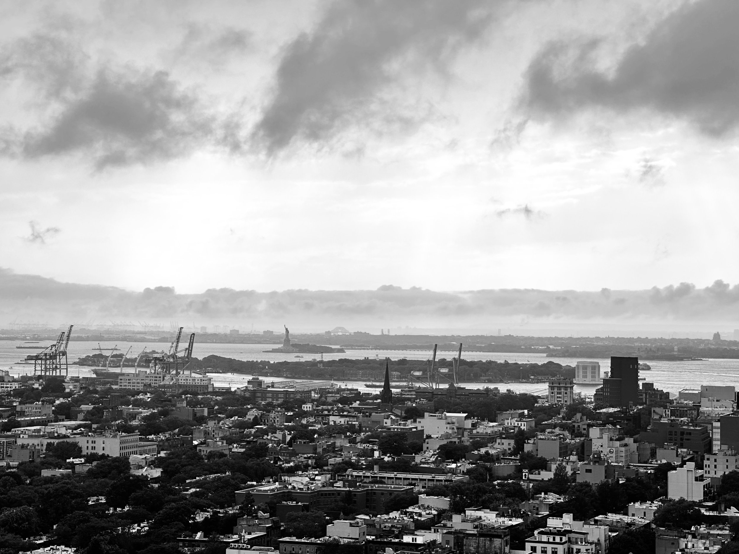 Black and white cityscape view of New York City, including the Statue of Liberty and the harbor with cranes, skyline, and cloudy sky.