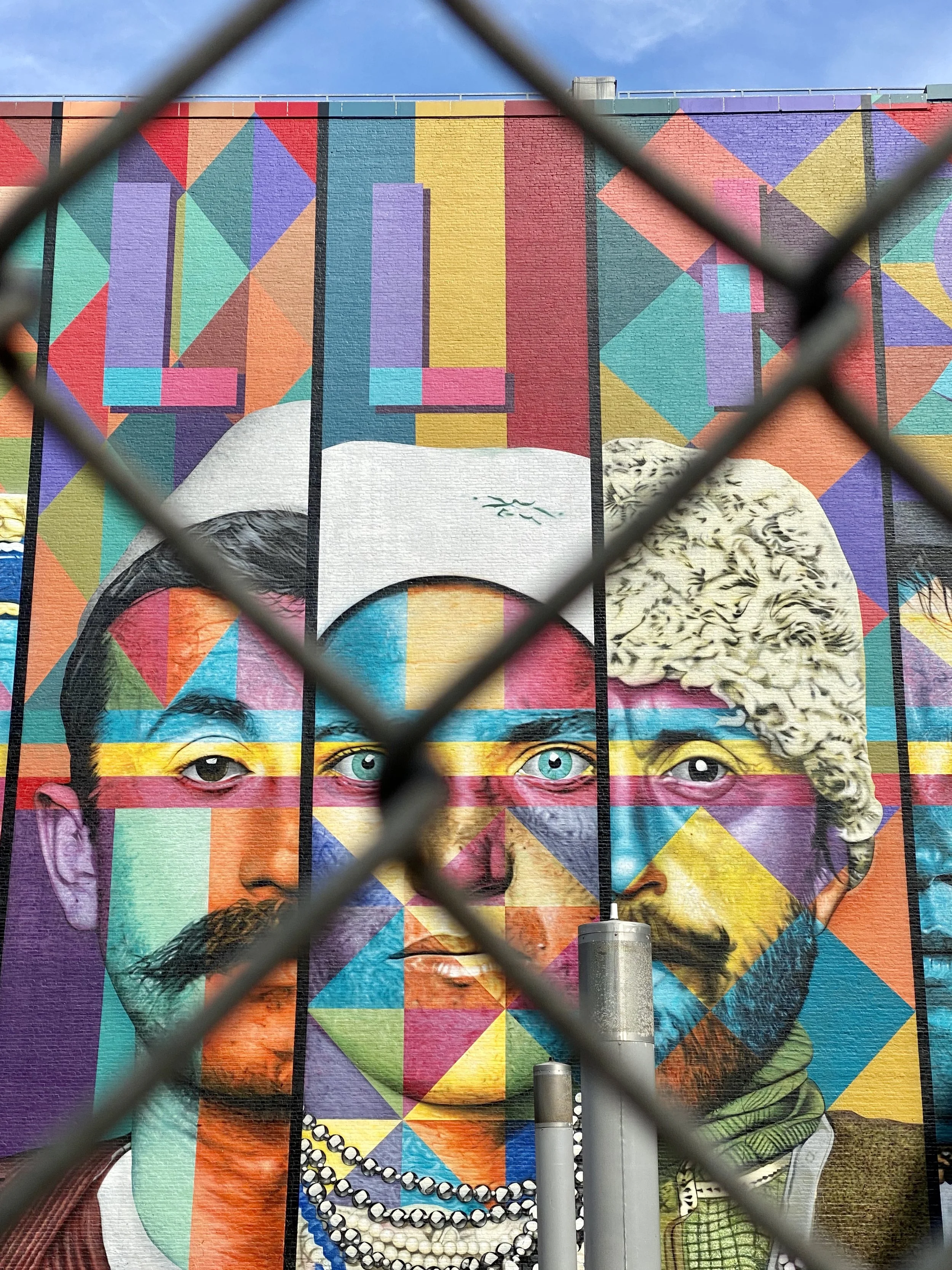 Colorful mural depicting a historical figure with a beard, wearing a tudor-style outfit, depicted in a geometric style with bright colors, behind a chain-link fence.