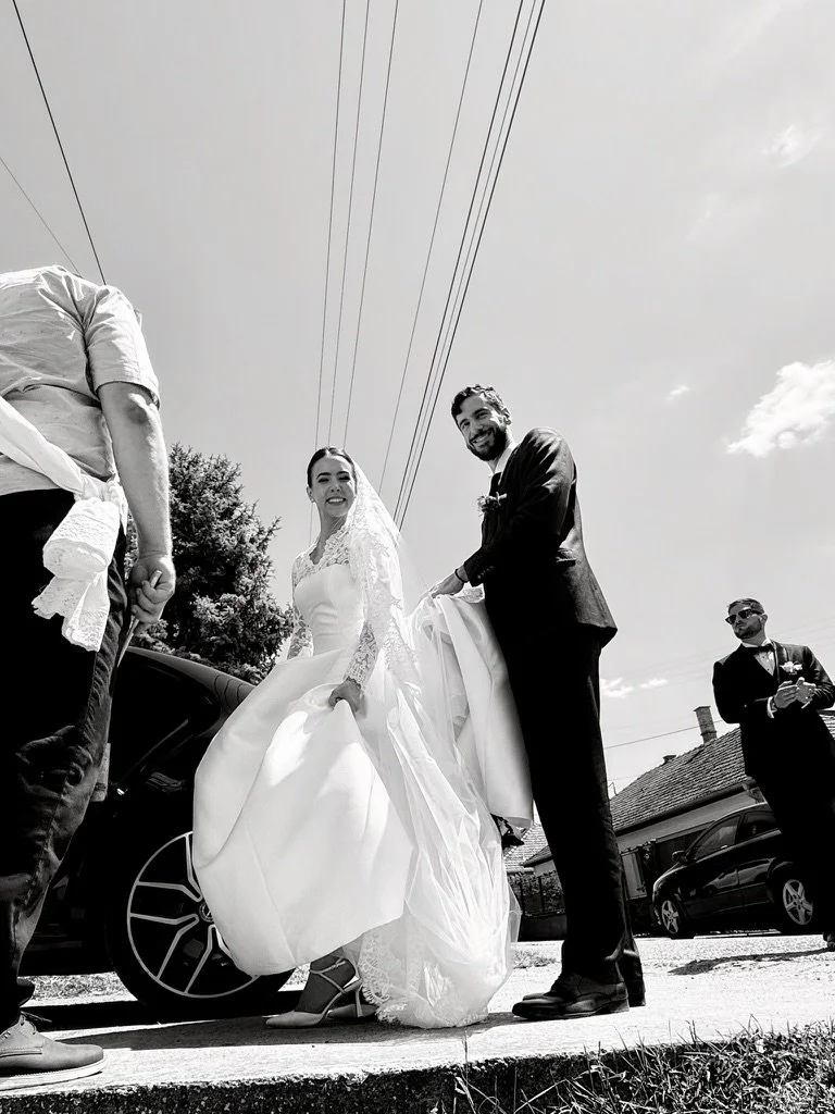 Bride in a white wedding dress holding up her gown, smiling, with a groom in a dark suit and tie beside her, outdoors with cars and houses in the background.