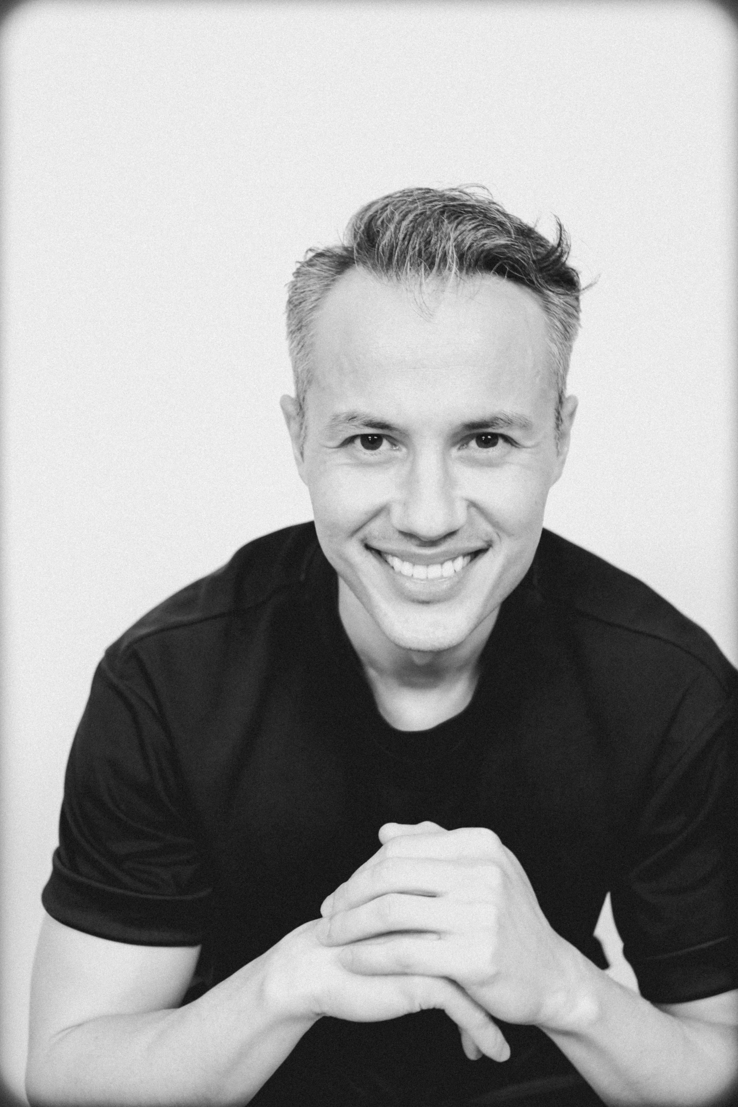 Black and white photo of a smiling man with combed hair, wearing a black shirt. He is resting his chin on his clasped hands and looking at the camera.