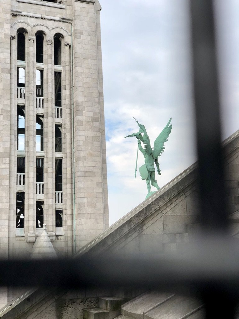 View of a historical building with a large clock tower and a green statue of a winged figure on a sloped roof.