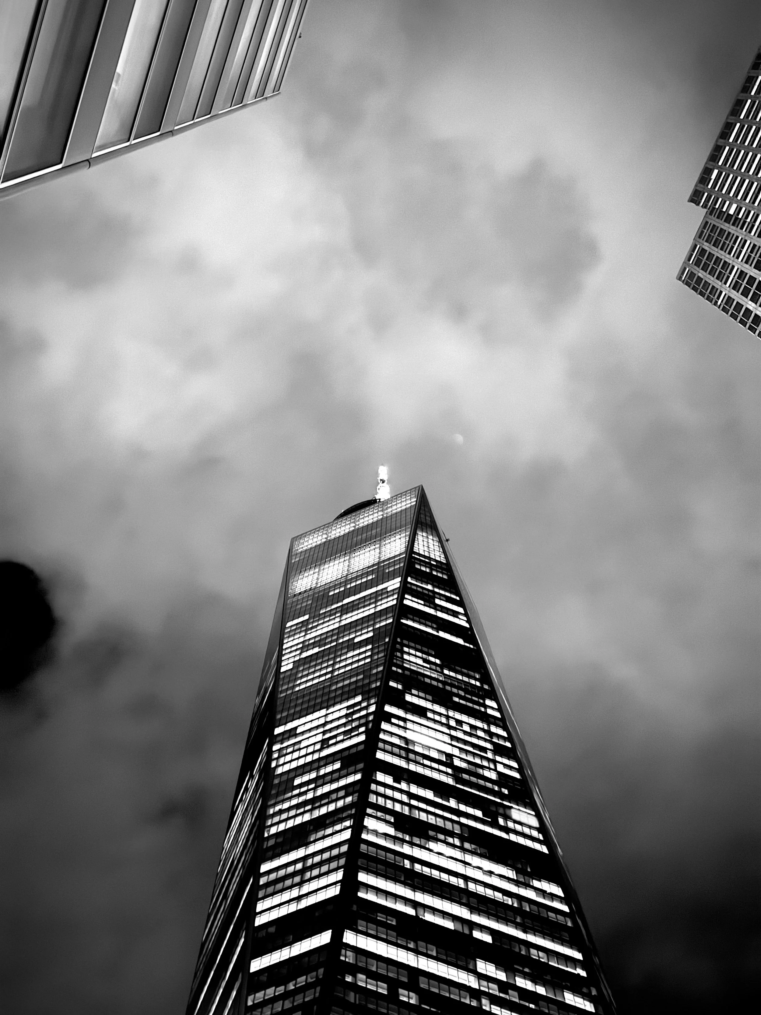 Black-and-white photo of a towering skyscraper viewed from below, with neighboring buildings and a cloudy sky.