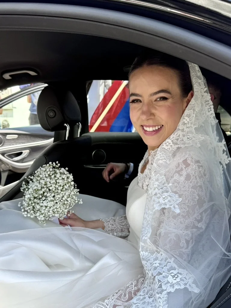 A bride sitting in a car, smiling, wearing a white wedding dress and veil, holding a bouquet of white baby's breath flowers.